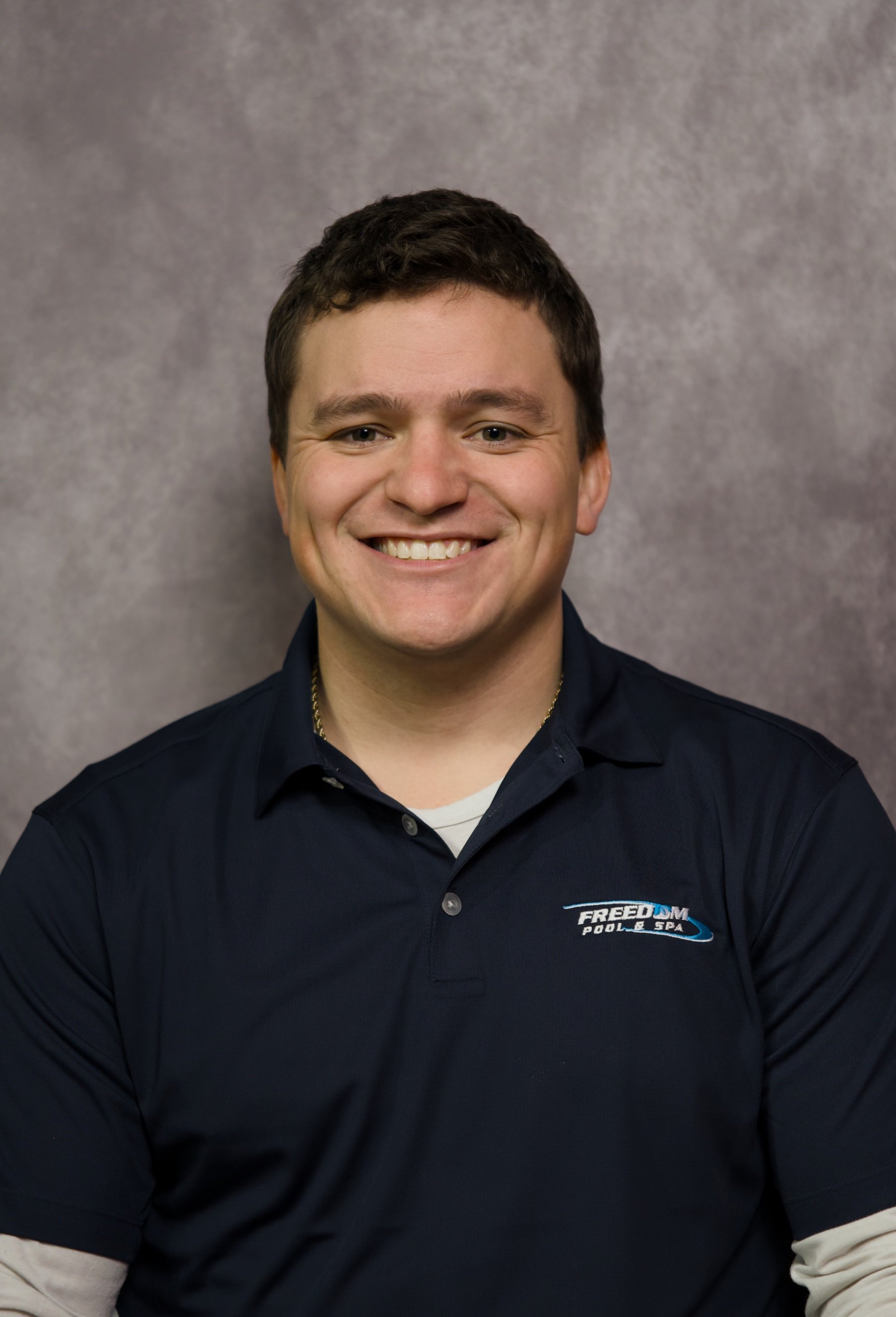 A smiling man with short brown hair wearing a navy blue FREDOM Pool & Spa polo shirt, standing against a gray background.
