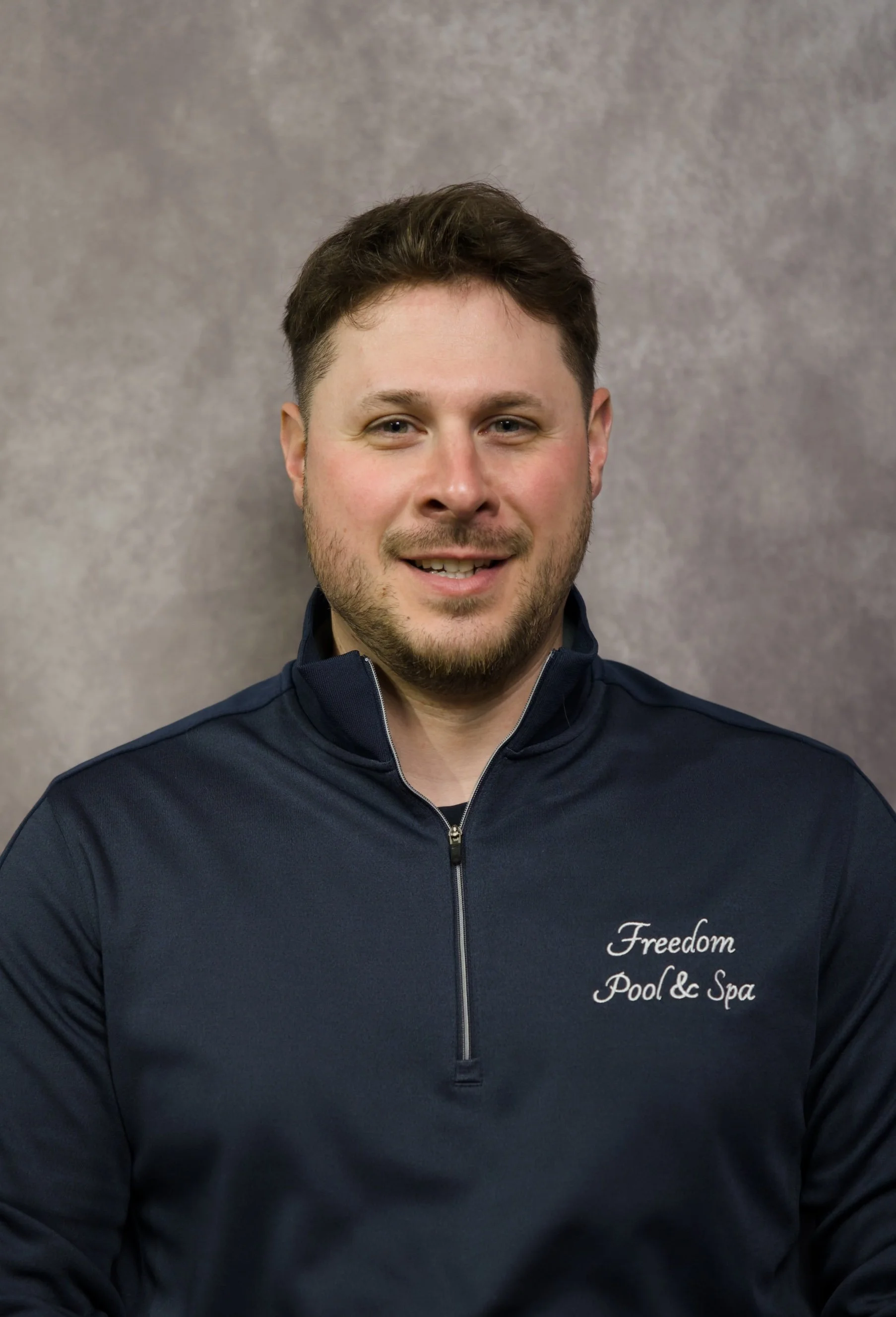 Portrait of a man in a navy jacket with the text 'Freedom Pool & Spa' on it, smiling at the camera, against a neutral background.