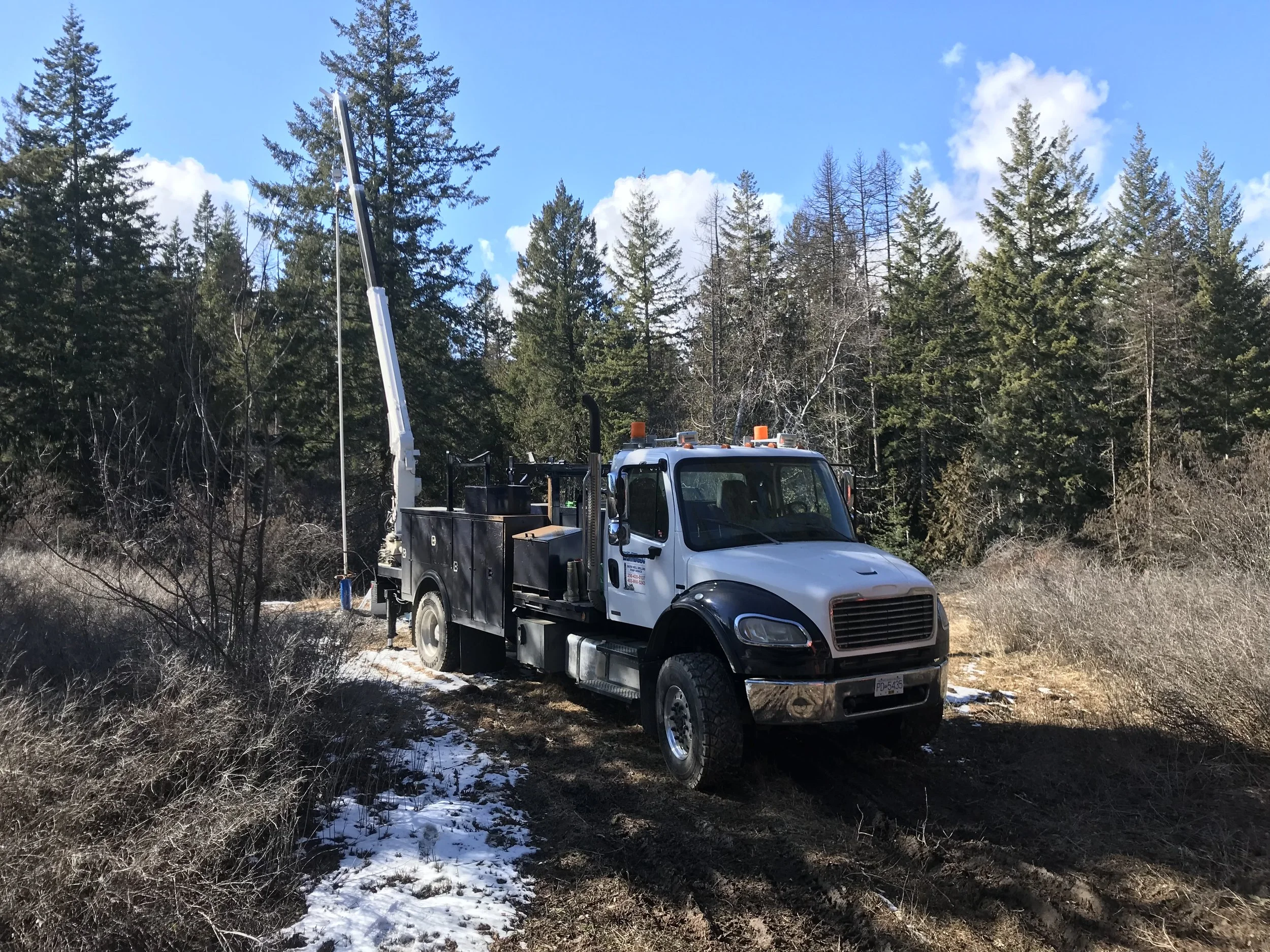 A utility truck with a crane arm and orange warning lights in a wooded area with tall trees, partially snow-covered ground, and a bright blue sky.
