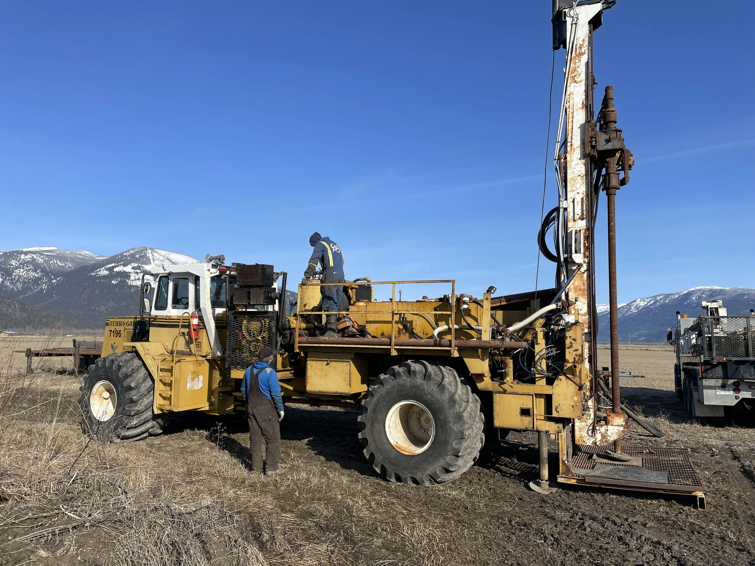 Two workers operating a large yellow drilling machine in an open field with mountains in the background and a clear blue sky.