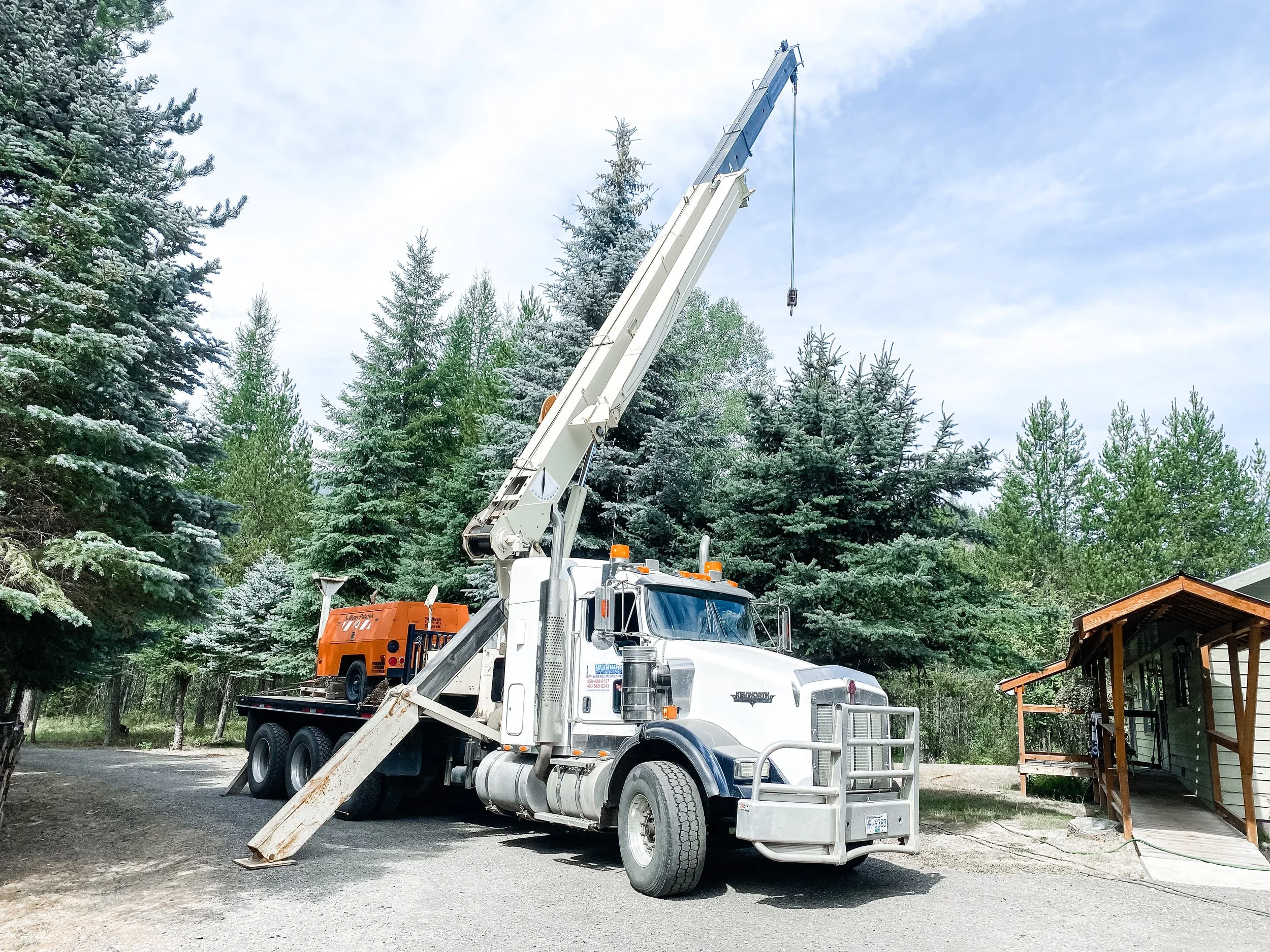 A large white tow truck with an extended crane arm parked on a dirt ground next to a green house. The background features a forest of pine trees under a cloudy sky.