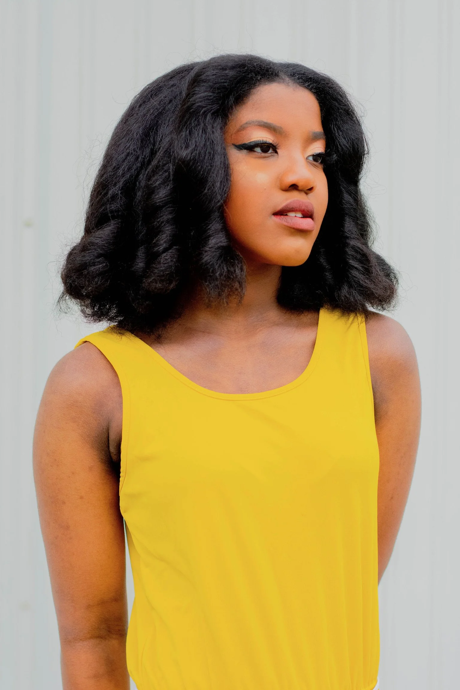 A young woman with curly black hair wearing a sleeveless yellow top standing against a plain background.