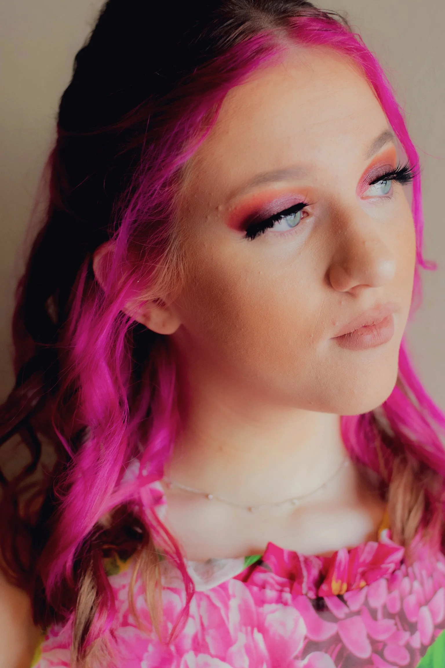 A woman with pink and brown hair, styled in loose waves, wearing pink eyeshadow, long black eyelashes, a floral dress with pink, purple, and green colors, and a delicate necklace. She looks to the side with a neutral expression.