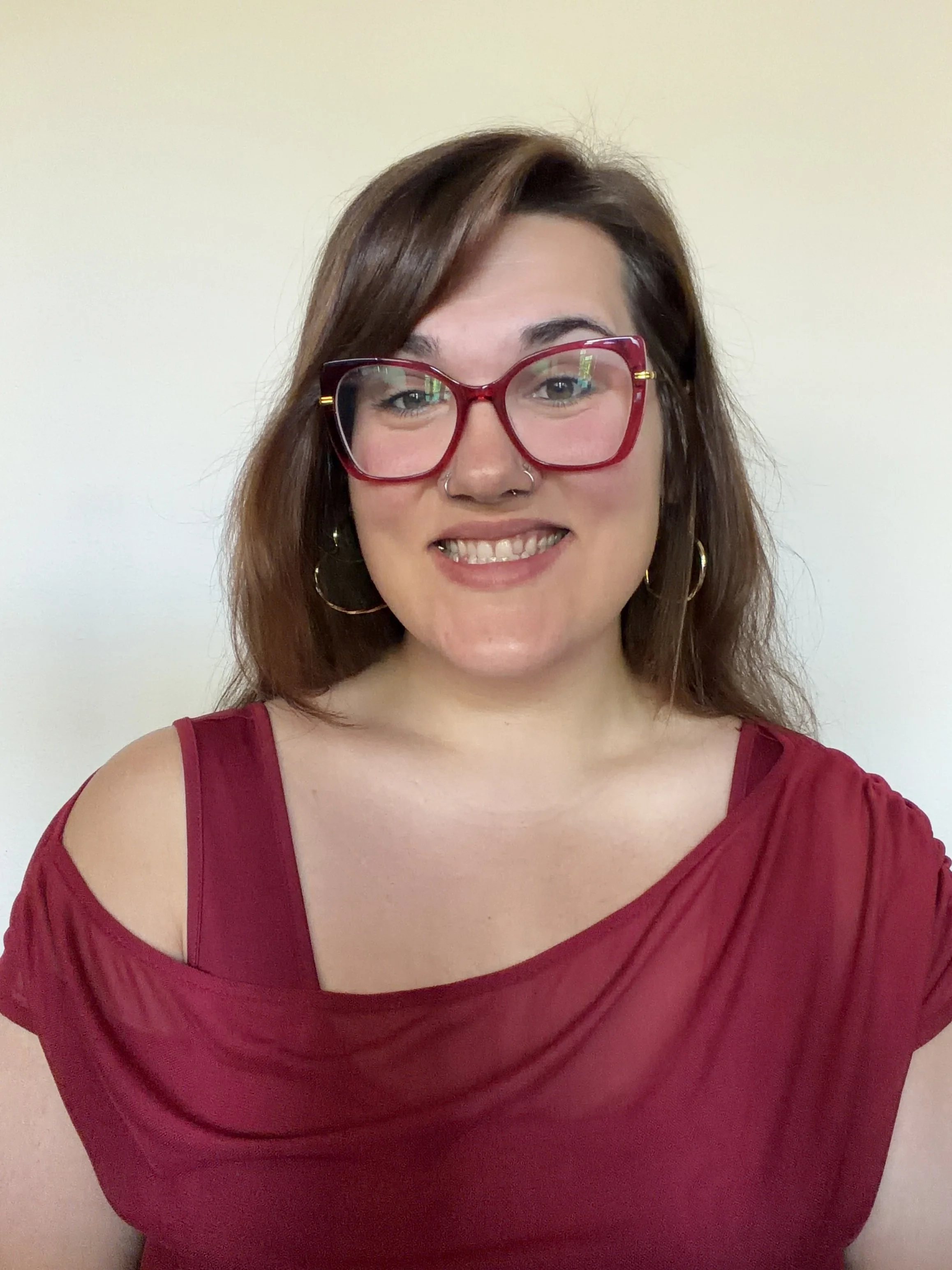A woman smiling wearing a red dress and red glasses, with shoulder-length brown hair, a nose ring, and hoop earrings, standing against a plain, light-colored wall.