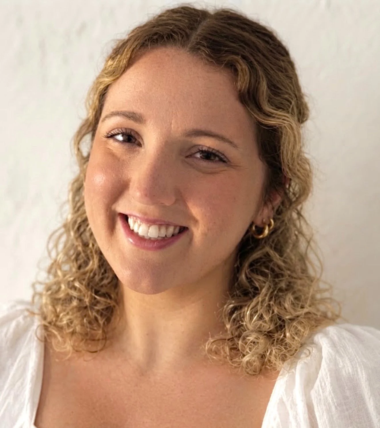 Close-up of a woman with curly blonde hair, wearing a white top and gold hoop earrings, smiling at the camera against a plain white background.