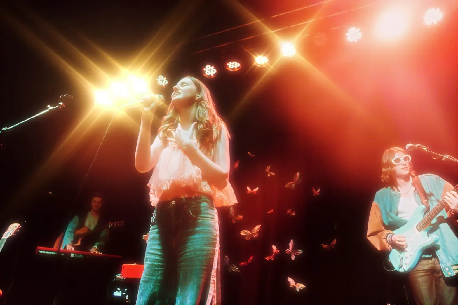 A musical performance with a female singer and band members on stage, with backlighting and butterfly visuals.