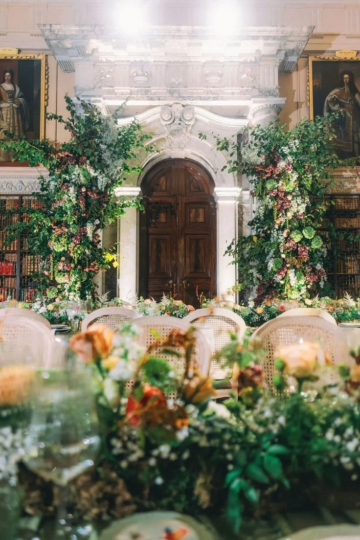 Long banquet table in a grand historic hall with green-draped tables, elegant place settings and lush floral installations creating a dramatic, symmetrical dining experience.