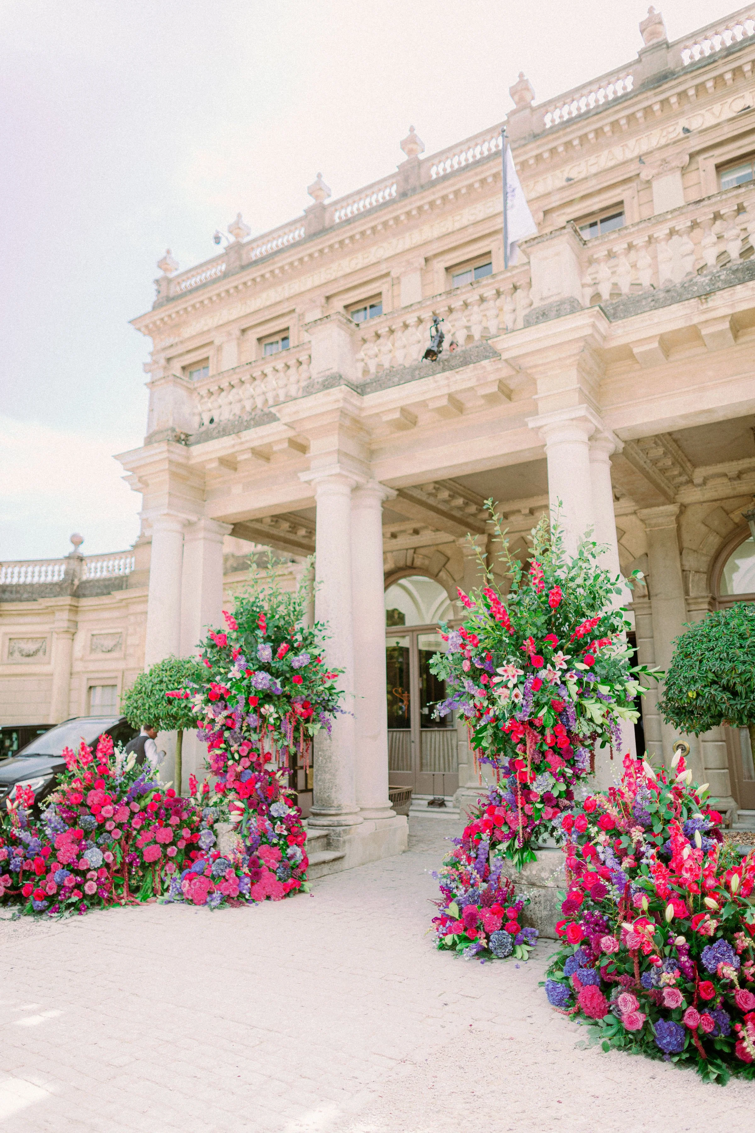 Grand neoclassical building exterior adorned with abundant floral arrangements in pinks, purples and greens framing the entrance for a luxurious celebration.