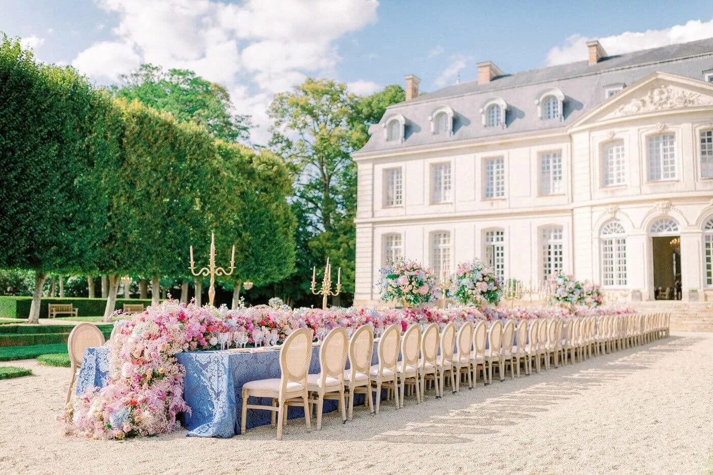 An elegant outdoor banquet table adorned with pink and purple flowers and gold candelabras stretches in front of a grand white chateau on a sunny day. Classic chairs are arranged along each side of the table.