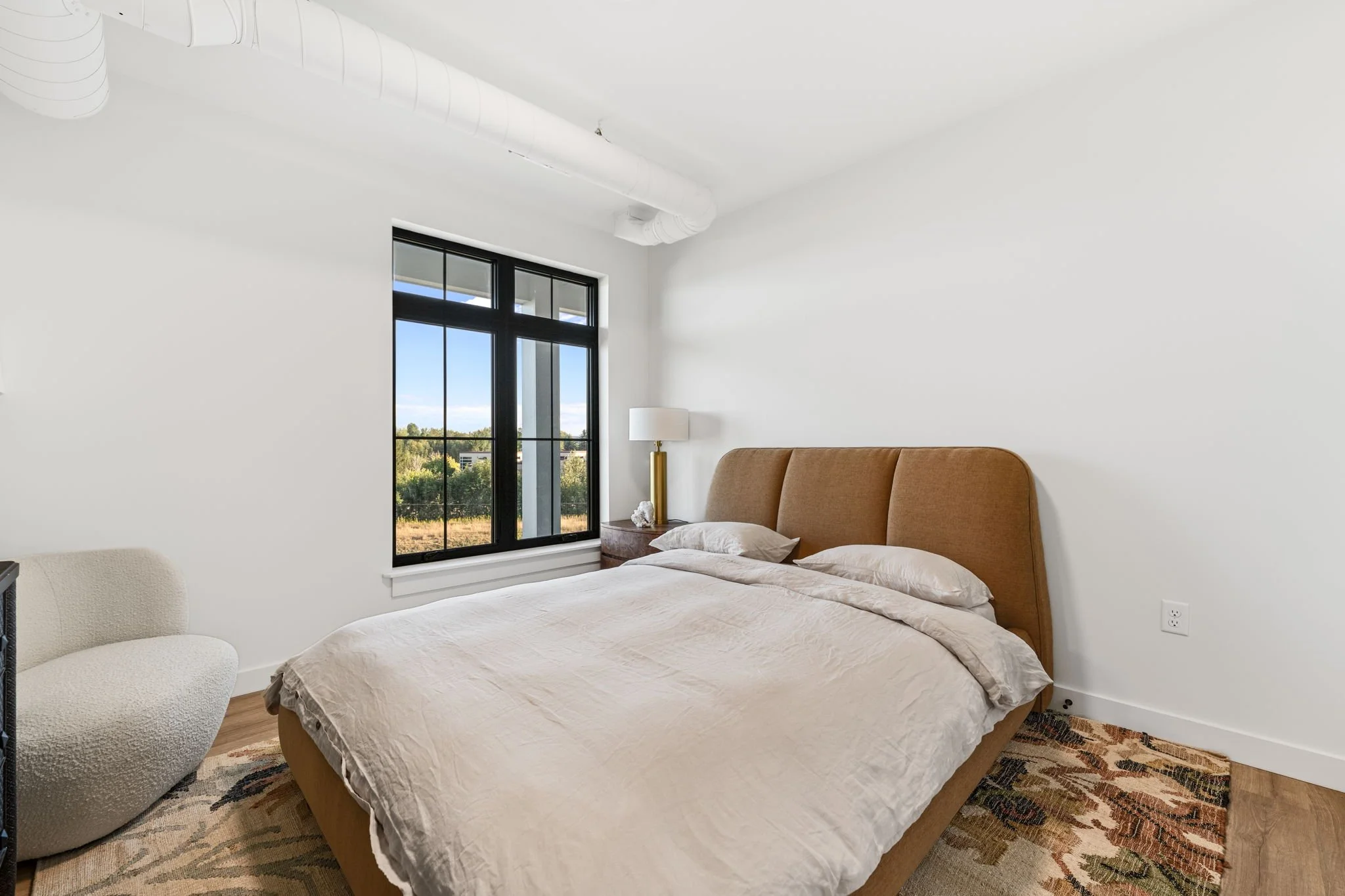 A bedroom with a bed featuring a tan headboard, white bedding, and pillows. A window with black frames shows a view of a green outdoor area. The Residences at North Harbor on Lake Superior in Marquette Michigan.