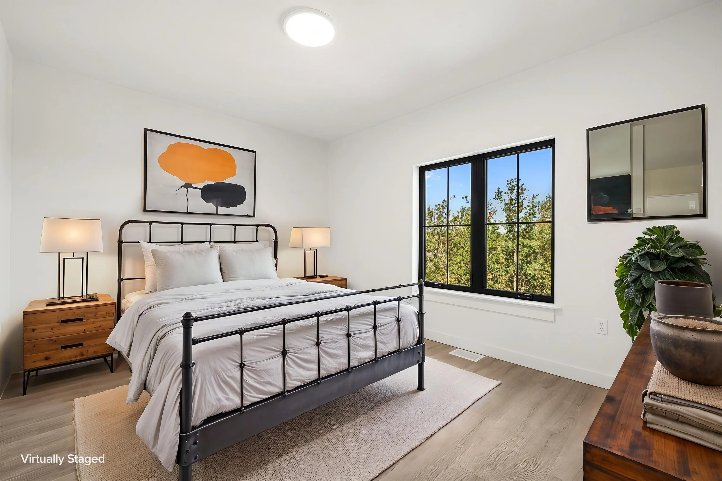 A bedroom with a black metal bed frame, white bedding, two bedside wooden tables with lamps, a large window showing trees. The Residences at North Harbor on Lake Superior in Marquette Michigan.