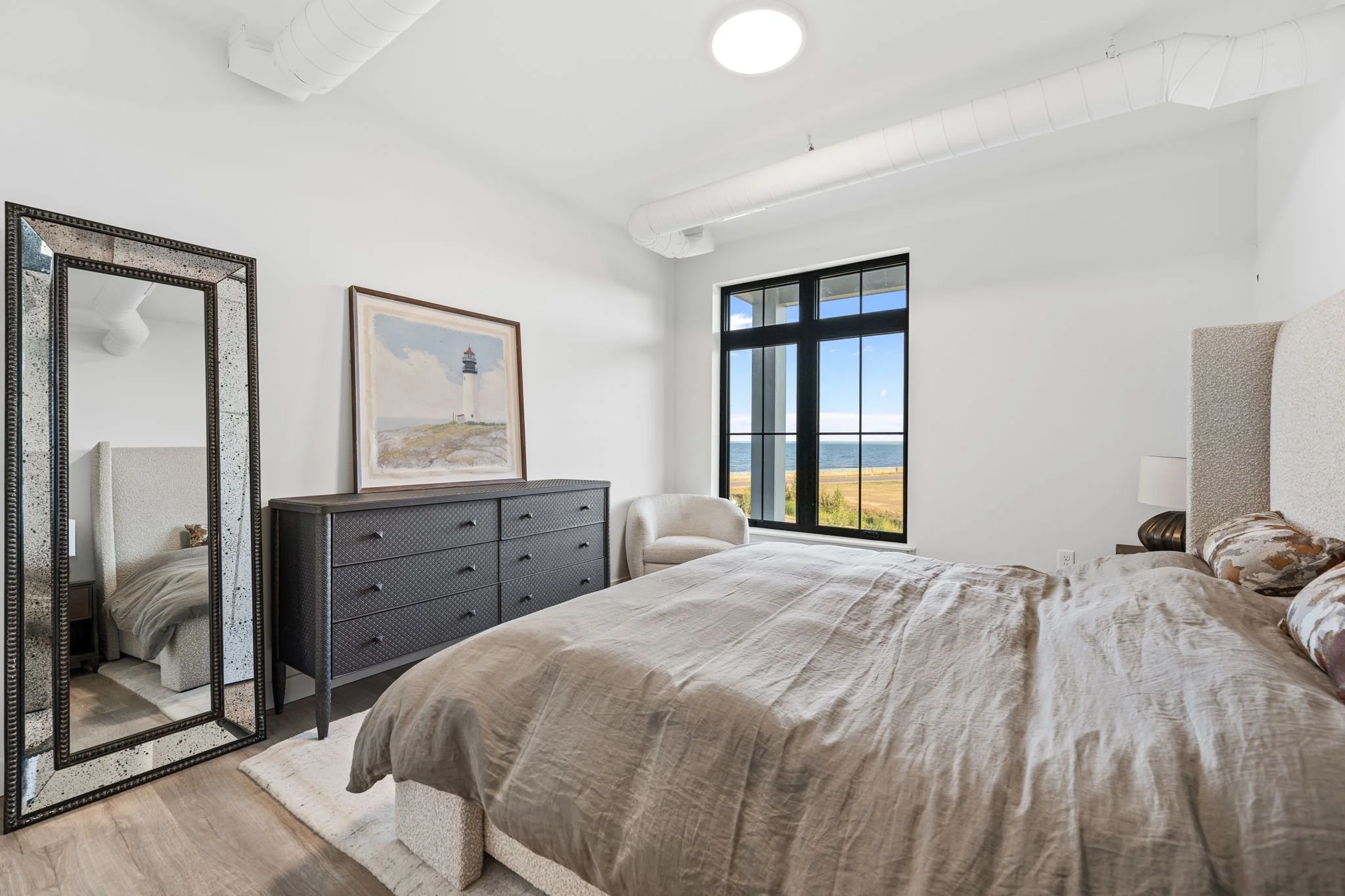 A modern bedroom with white walls and a large window showing an outdoor view of the ocean and clear sky. The Residences at North Harbor on Lake Superior in Marquette Michigan.