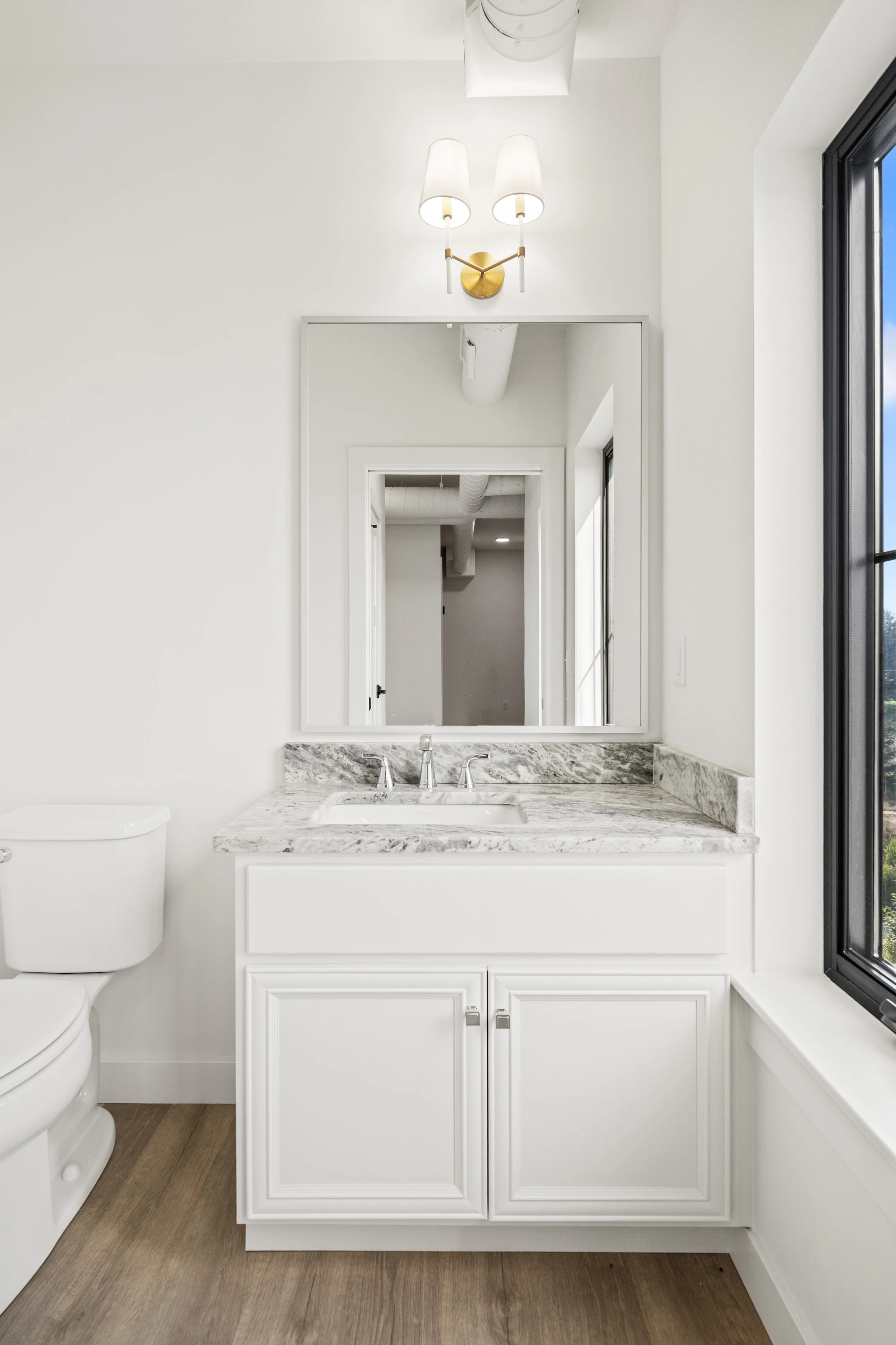 Bathroom with white vanity, marble countertop, mirror, wall-mounted light fixture, window, and toilet. The Residences at North Harbor on Lake Superior in Marquette Michigan.