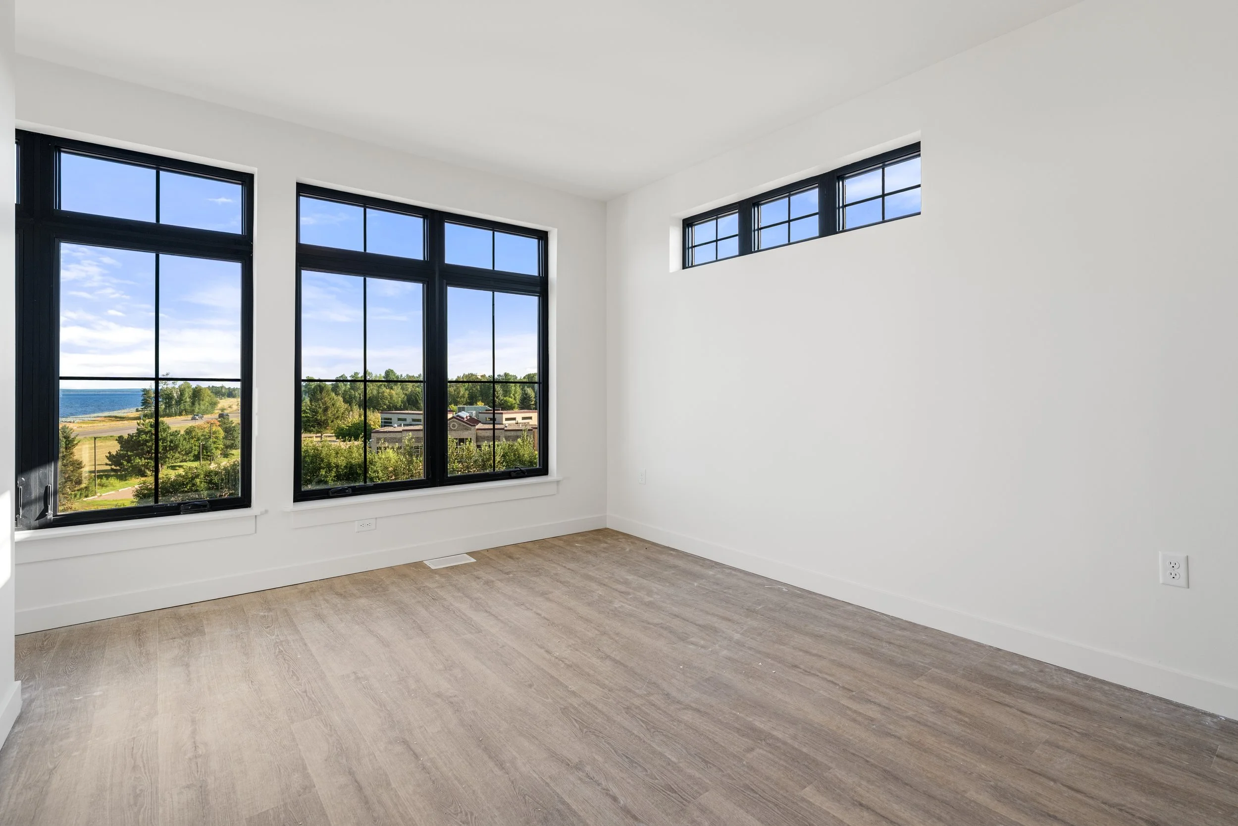 Empty room with large black-framed windows showing a view of trees, buildings, and a body of water under a blue sky with clouds. The Residences at North Harbor on Lake Superior in Marquette Michigan.