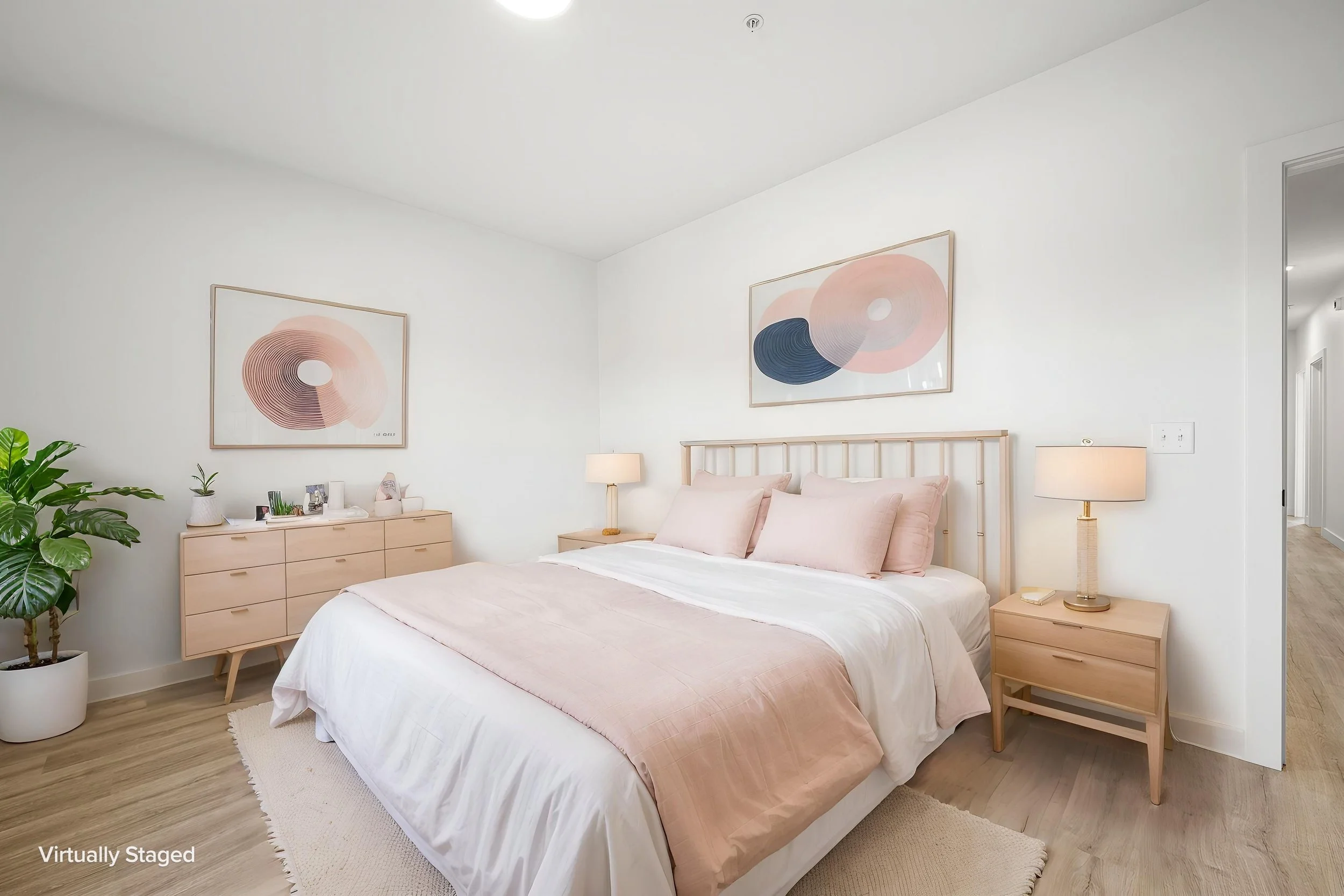 A bedroom with a bed, two nightstands with lamps, a dresser, and framed abstract art on the wall. There is a green potted plant on the left and a hallway on the right. The Residences at North Harbor on Lake Superior in Marquette Michigan.