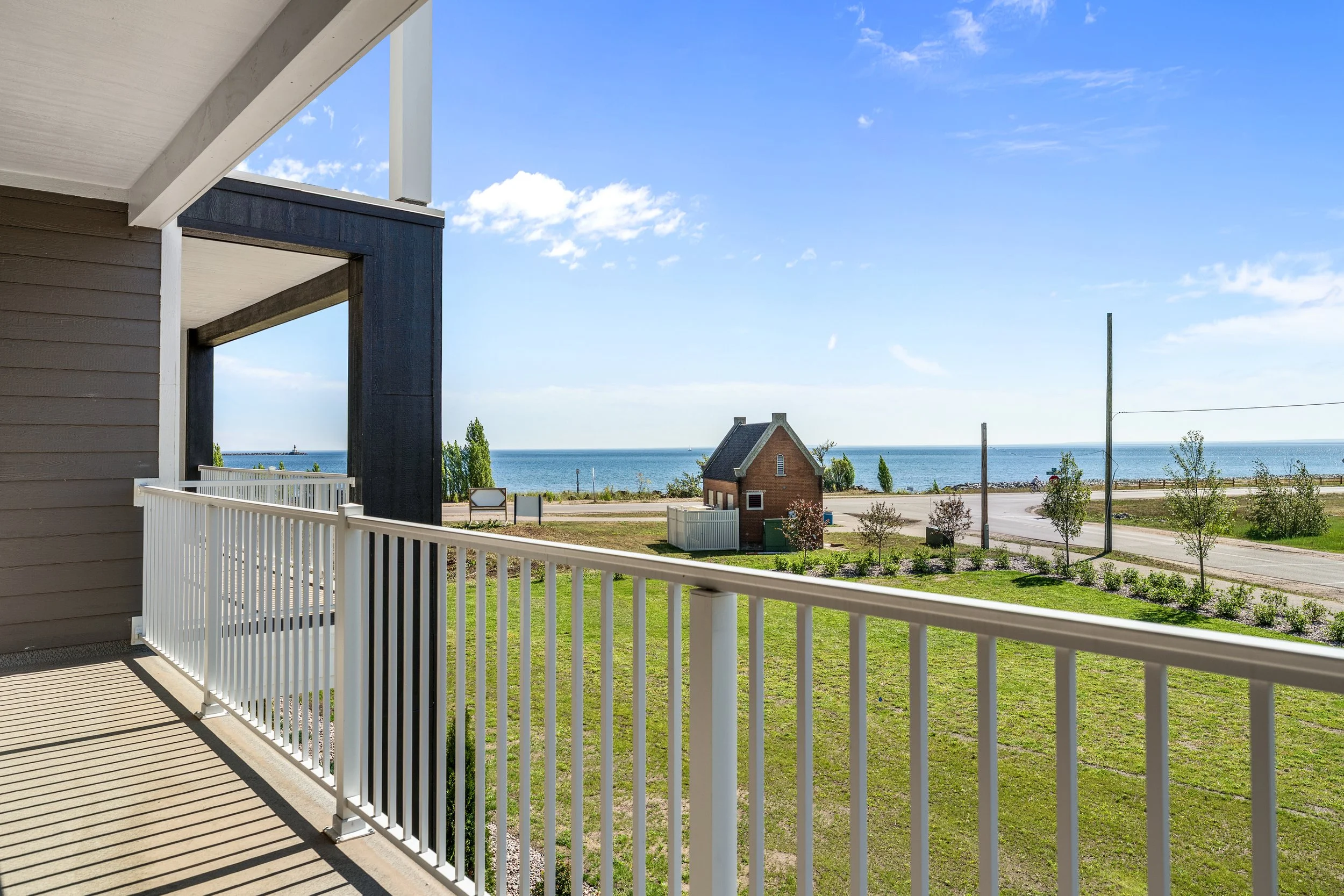 View from a balcony overlooking a grassy yard and the ocean with a clear blue sky. There is a small brick house and some young trees in the landscape. The Residences at North Harbor on Lake Superior in Marquette Michigan.
