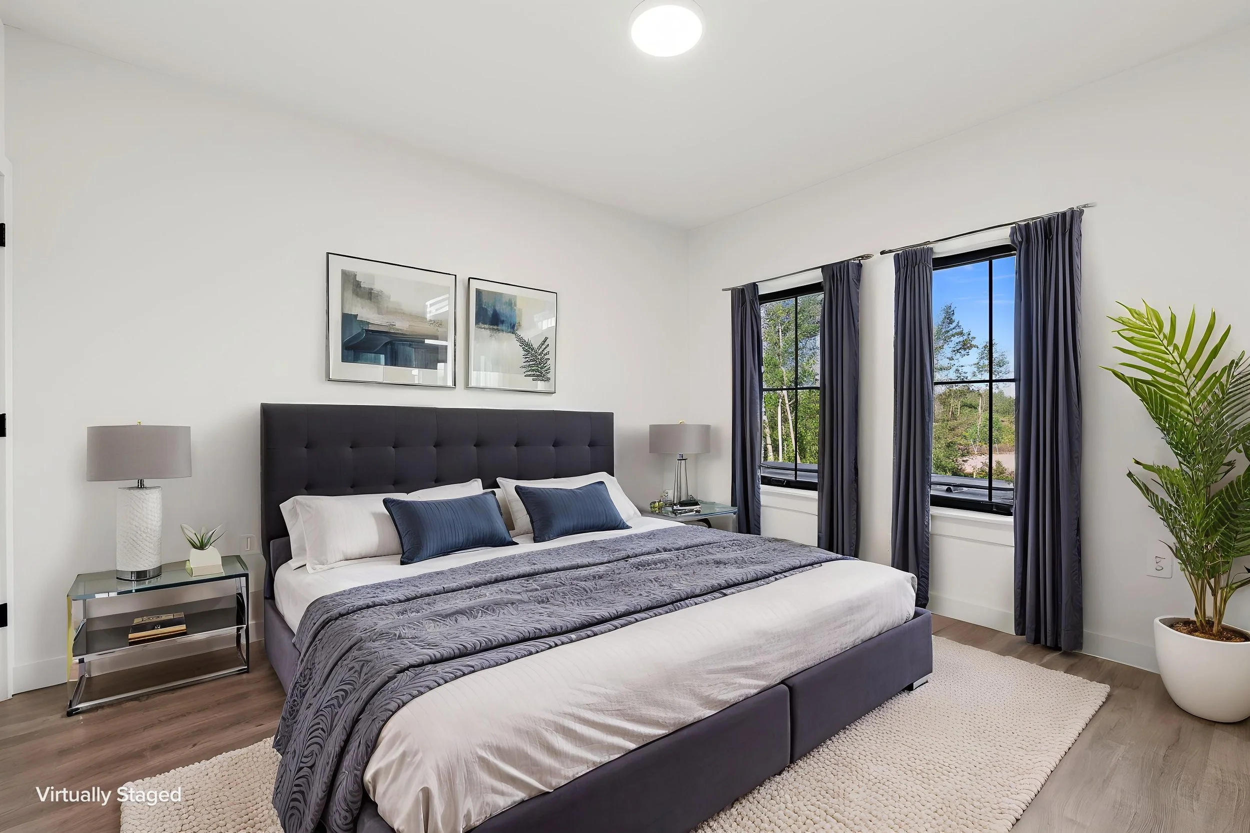 A modern bedroom with a large bed featuring a gray upholstered headboard, white sheets, and blue pillows. Flanking the bed are two nightstands with lamps, a small plant, and books. The Residences at North Harbor on Lake Superior in Marquette Michigan