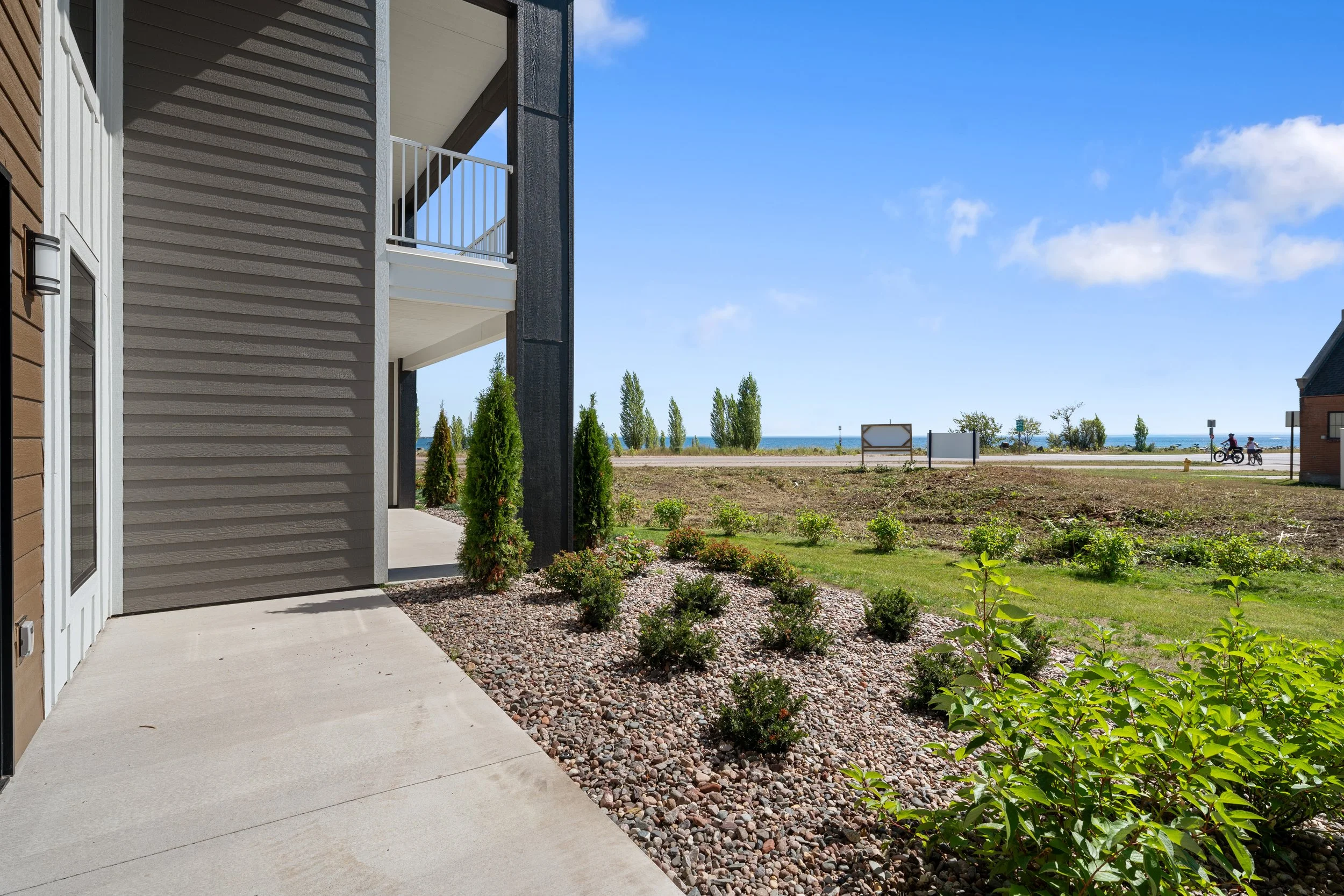 A building with gray horizontal siding and a balcony, next to a patch of landscaped plants. The Residences at North Harbor on Lake Superior in Marquette Michigan.