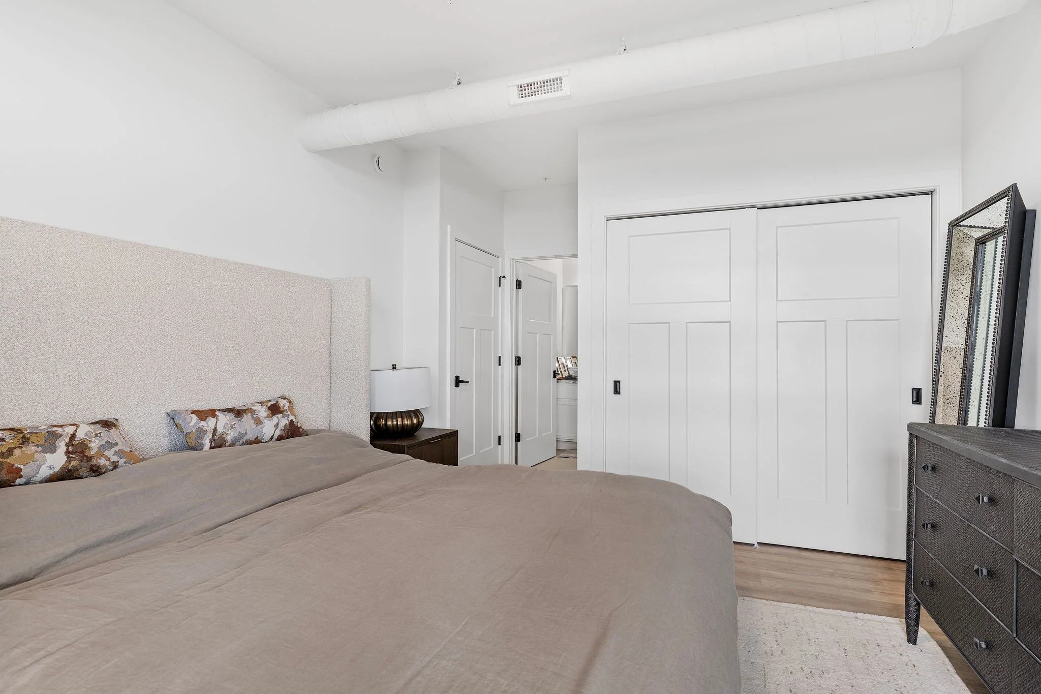 Bedroom with beige bedspread, floral pillows, white walls, a large mirror, a nightstand with a lamp, white closet doors, and a door leading to another room. The Residences at North Harbor on Lake Superior in Marquette Michigan.