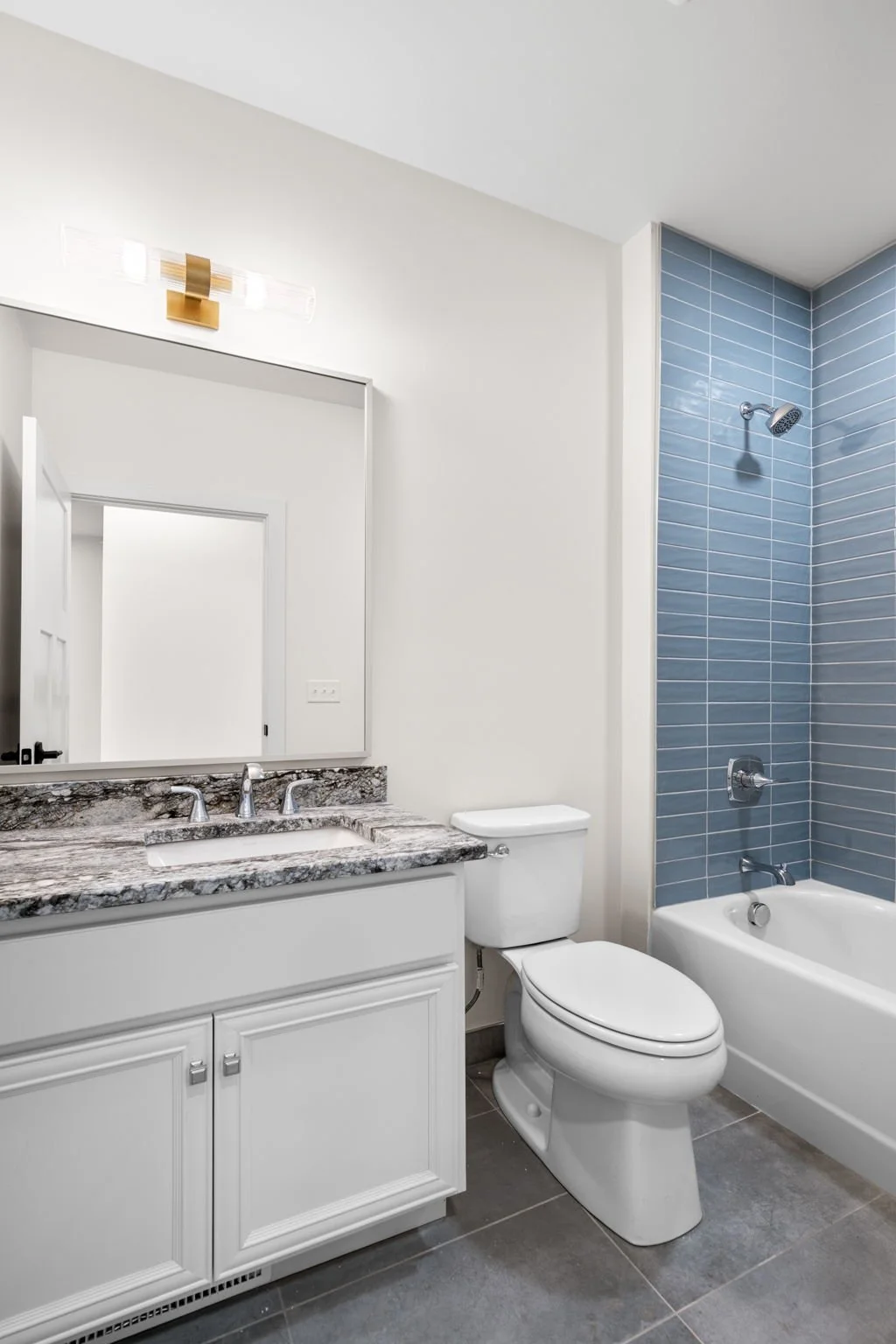Bathroom with white vanity, granite countertop, large mirror, white toilet, bathtub with blue tile wall, and modern light fixture. The Residences at North Harbor on Lake Superior in Marquette Michigan.