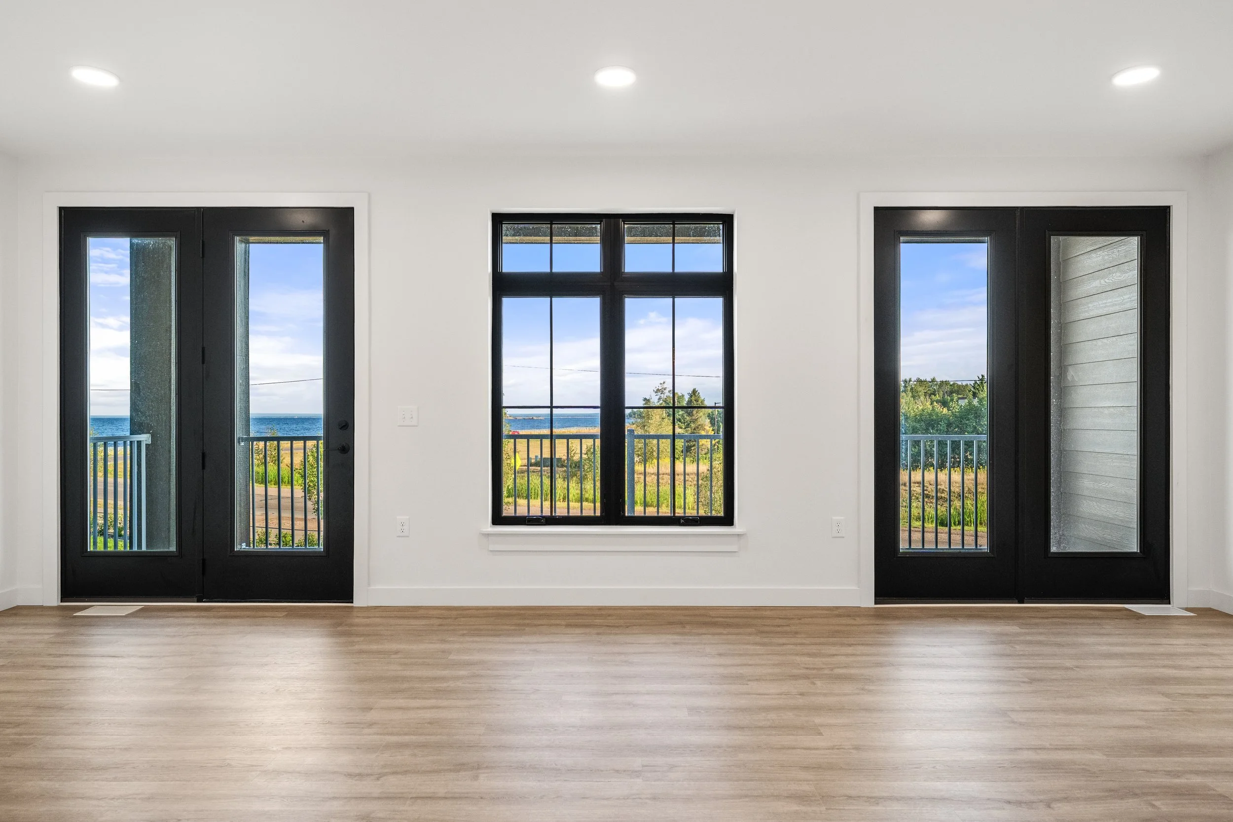 Interior view of a room with three black-framed windows showing an outdoor scenery including a body of water, trees, and a partly cloudy sky; plain white walls and light hardwood flooring. The Residences at North Harbor on Lake Superior in Marquette 
