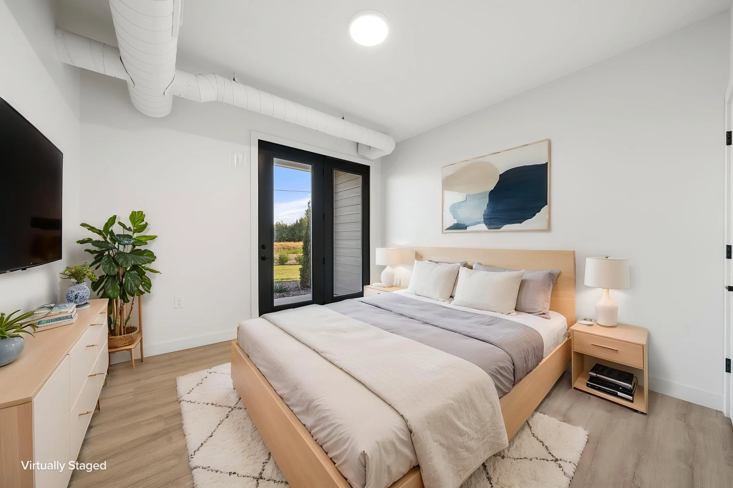 Modern bedroom with a wooden bed frame, light-colored bedding, a nightstand with a lamp, a piece of abstract wall art. The Residences at North Harbor on Lake Superior in Marquette Michigan.
