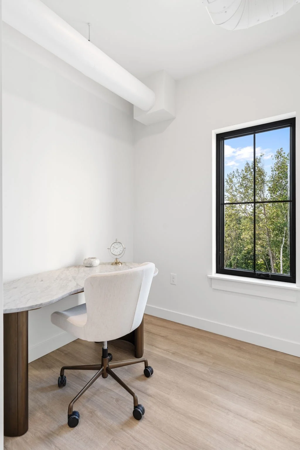 A minimalist home office with a white marble desk, white swivel chair, small clock on desk, and a large black-framed window showing trees and blue sky outside. The Residences at North Harbor on Lake Superior in Marquette Michigan.