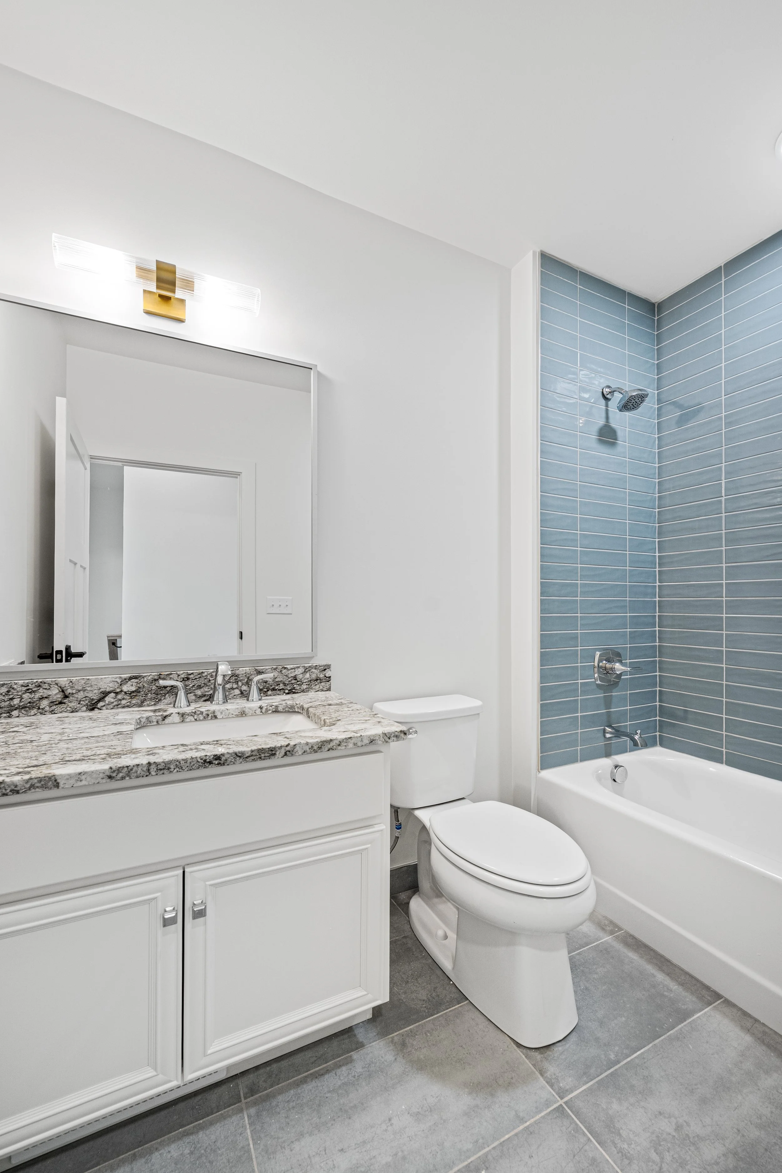 Modern bathroom featuring a white vanity with gray marble countertop, a large mirror, a white toilet, and a bathtub with blue tile surround and a showerhead. The Residences at North Harbor on Lake Superior in Marquette Michigan.