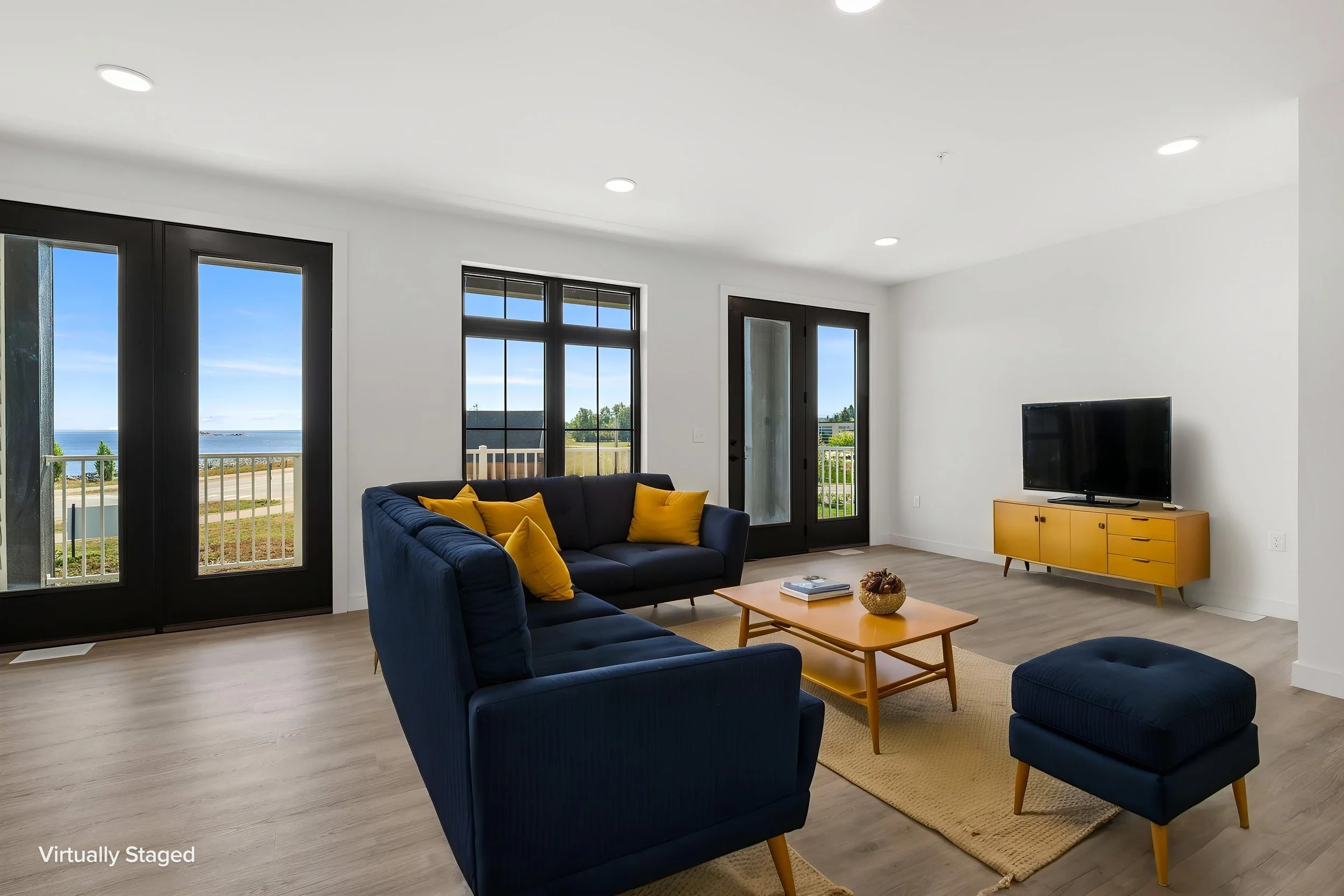 Living room with black-framed glass doors and windows showing outdoor view, navy blue sofas with yellow pillows, light wood coffee table, yellow TV stand, and wooden flooring. The Residences at North Harbor on Lake Superior in Marquette Michigan.