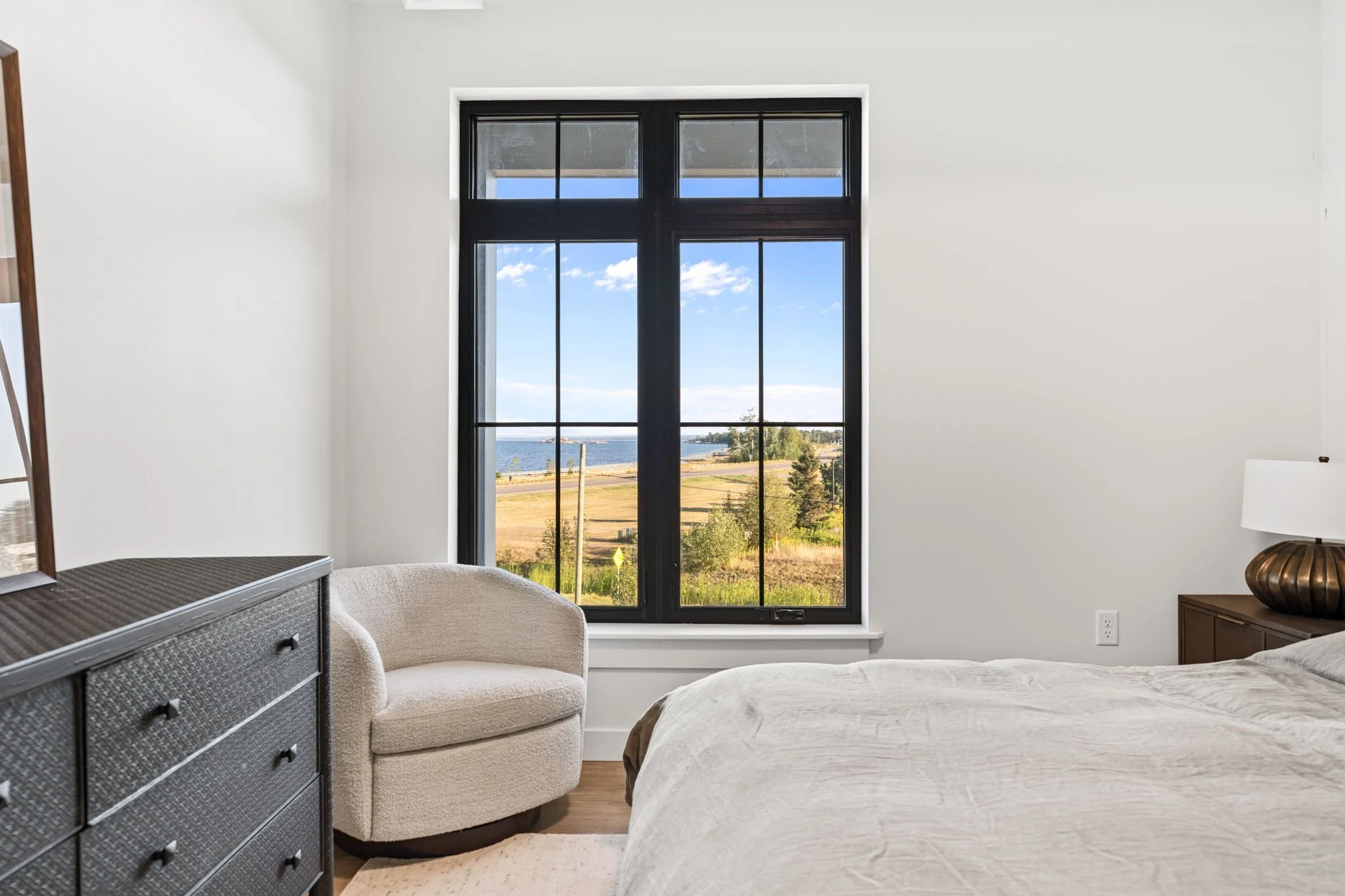 A bedroom with a large window showing a view of a blue sky, trees, and water outside. The room has a beige armchair, a dark gray dresser, and a nightstand with a decorative lamp. The Residences at North Harbor on Lake Superior in Marquette Michigan.