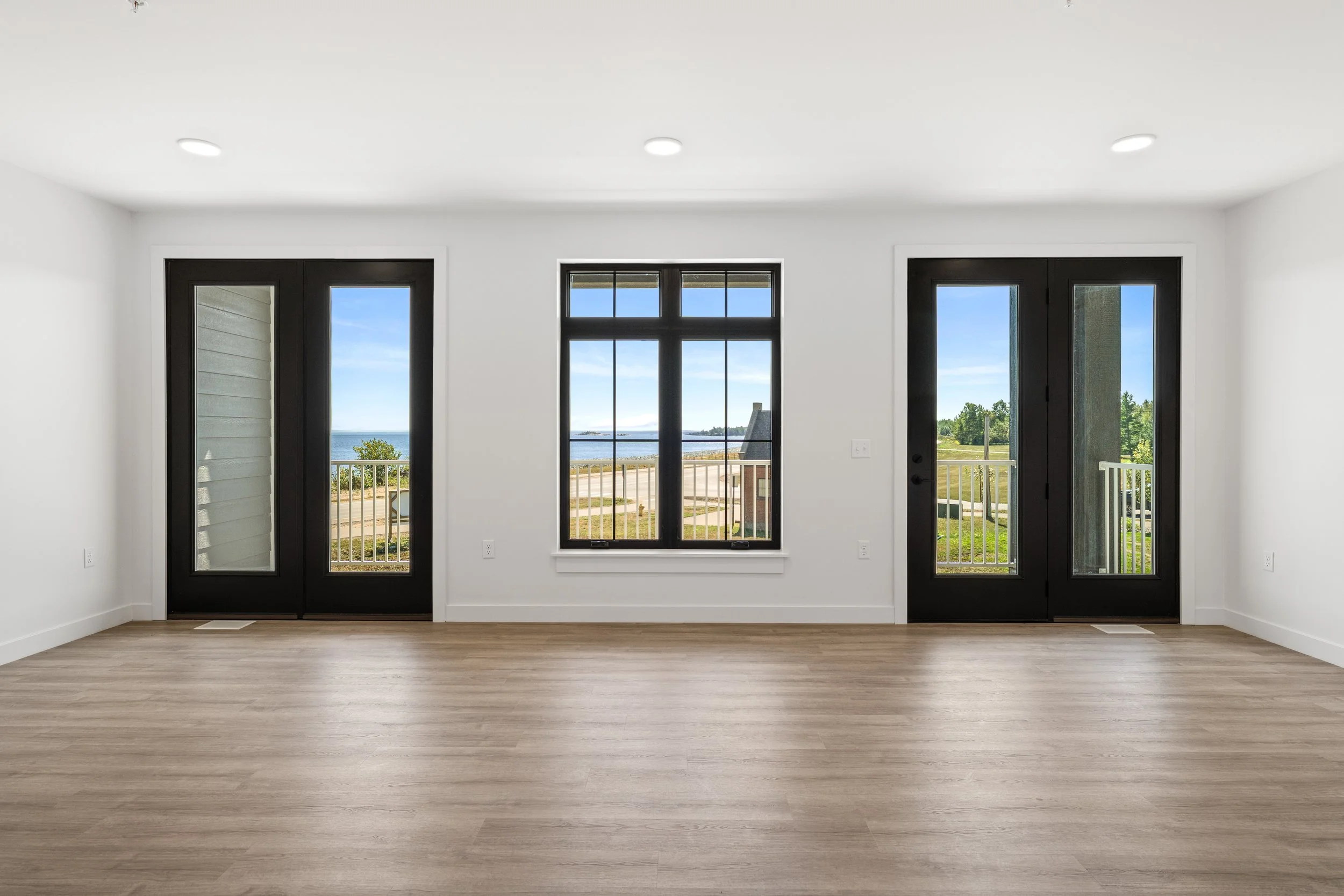 Room with white walls, light-colored wood flooring, three black framed glass doors, and a large window showing an outdoor view of water and greenery. The Residences at North Harbor on Lake Superior in Marquette Michigan.