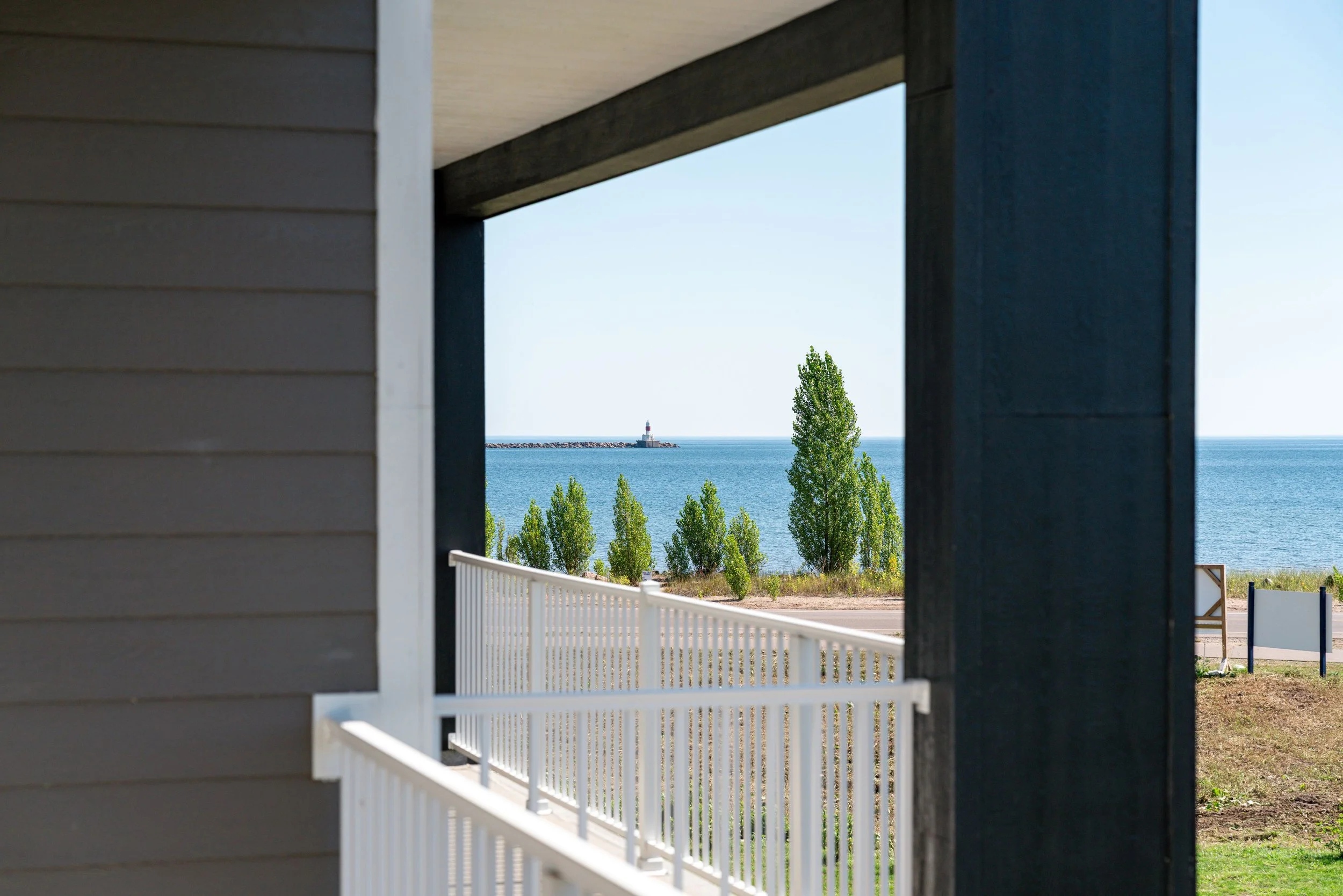 View of the ocean with a lighthouse on a breakwater, framed by part of a house or structure with horizontal siding and a white fence in the foreground. The Residences at North Harbor on Lake Superior in Marquette Michigan.
