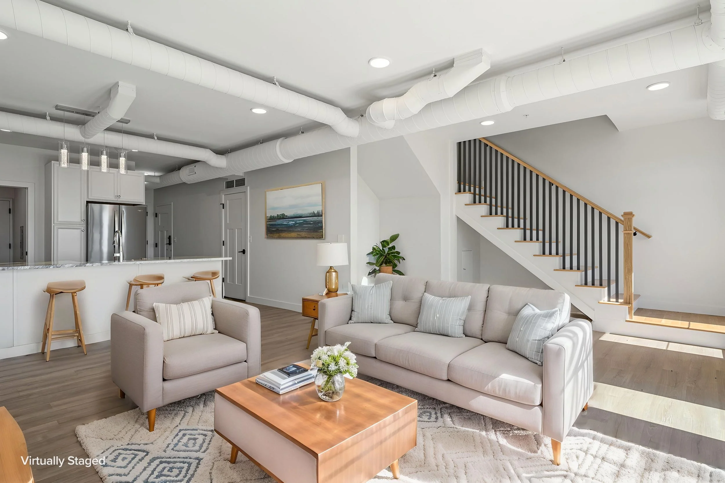 A modern open-concept living room with a beige sectional sofa, wooden coffee table with books and a flower arrangement, light-colored wooden flooring. The Residences at North Harbor on Lake Superior in Marquette Michigan.