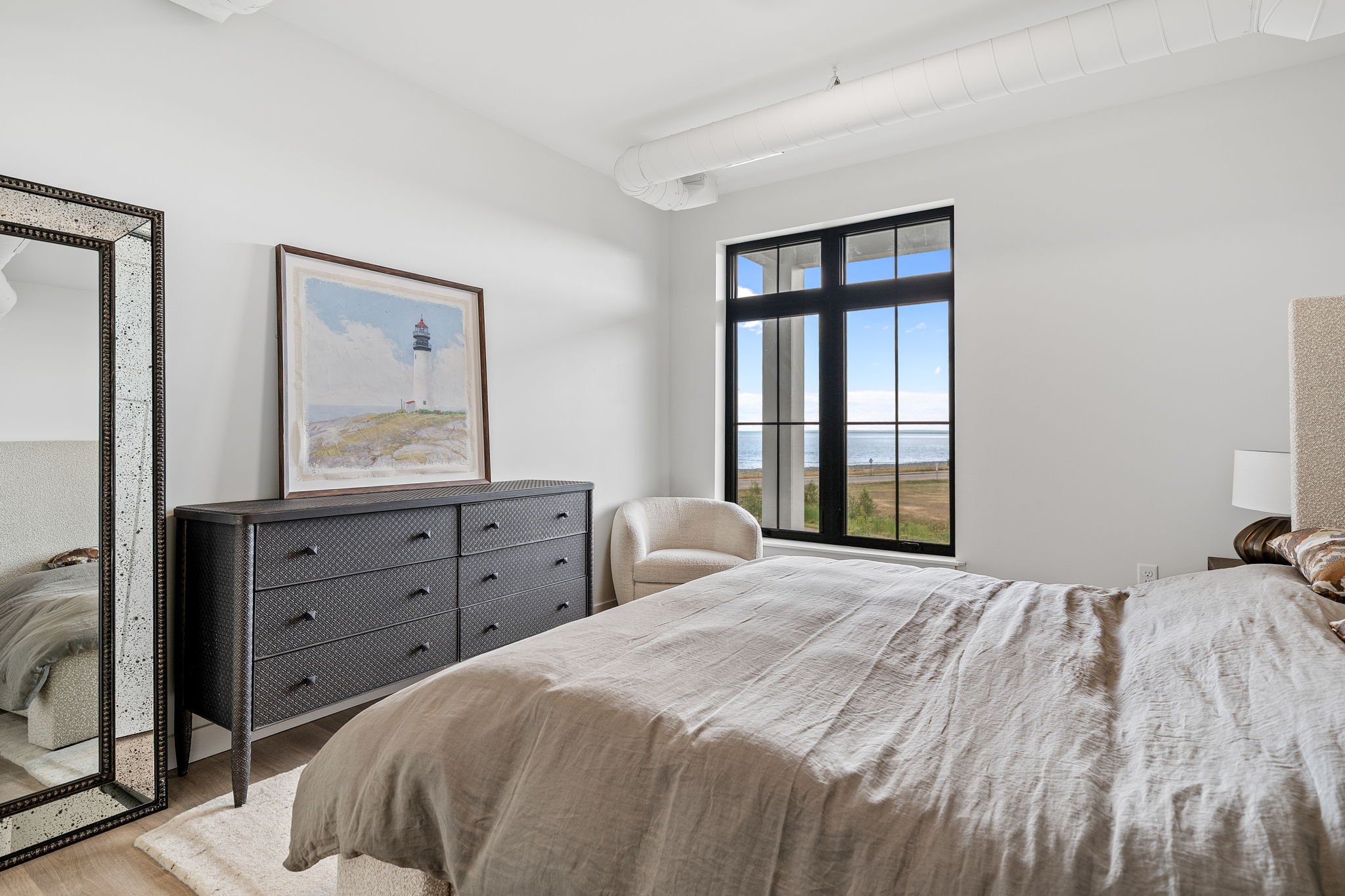 A bedroom with a large window showing a view of the outdoors, a black dresser with a framed painting of a lighthouse, a mirror, and an upholstered chair. The Residences at North Harbor on Lake Superior in Marquette Michigan.