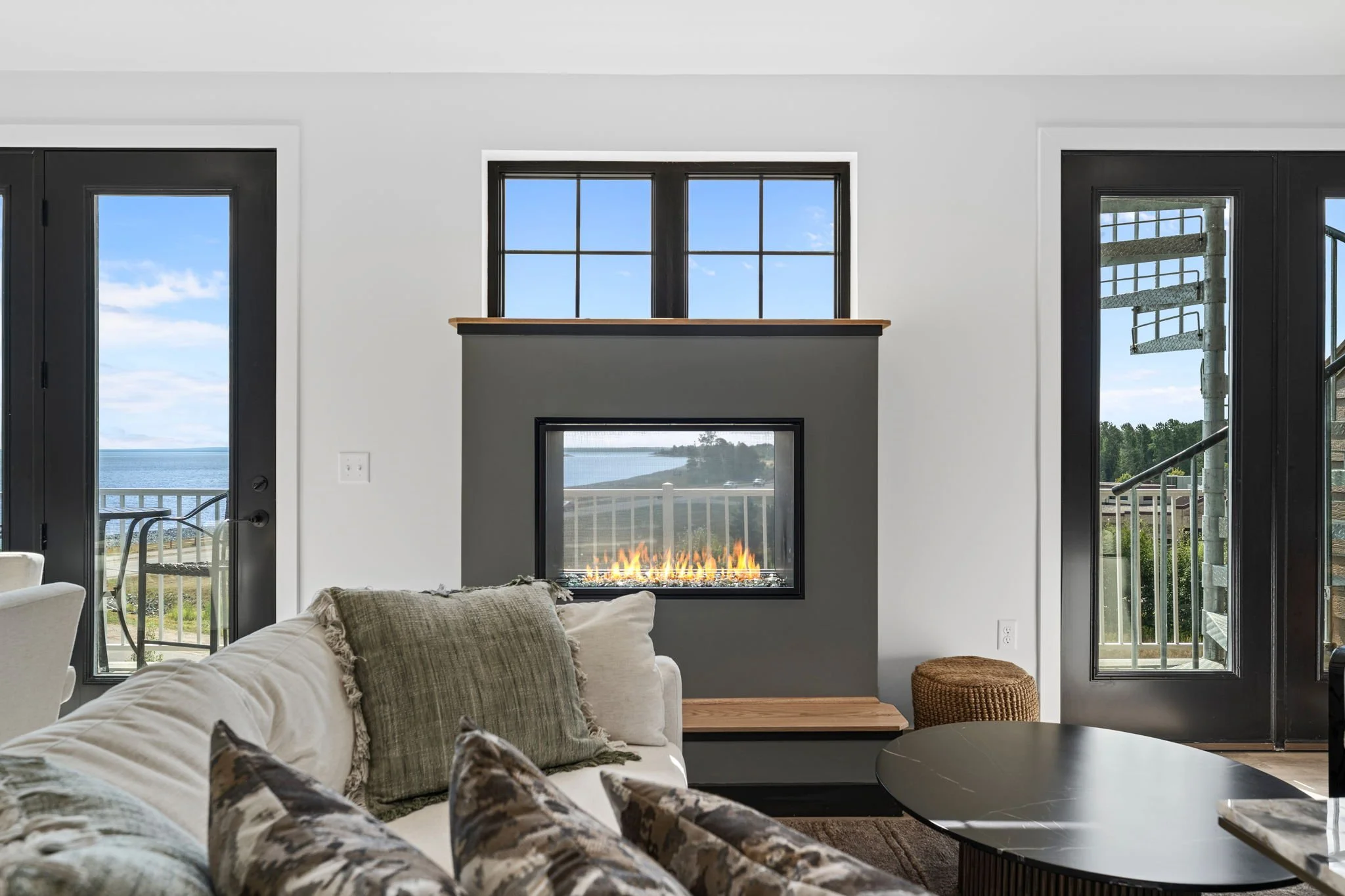 Living room with a fireplace, large windows showing a view of water and landscape, and a sectional sofa with patterned pillows. The Residences at North Harbor on Lake Superior in Marquette Michigan.