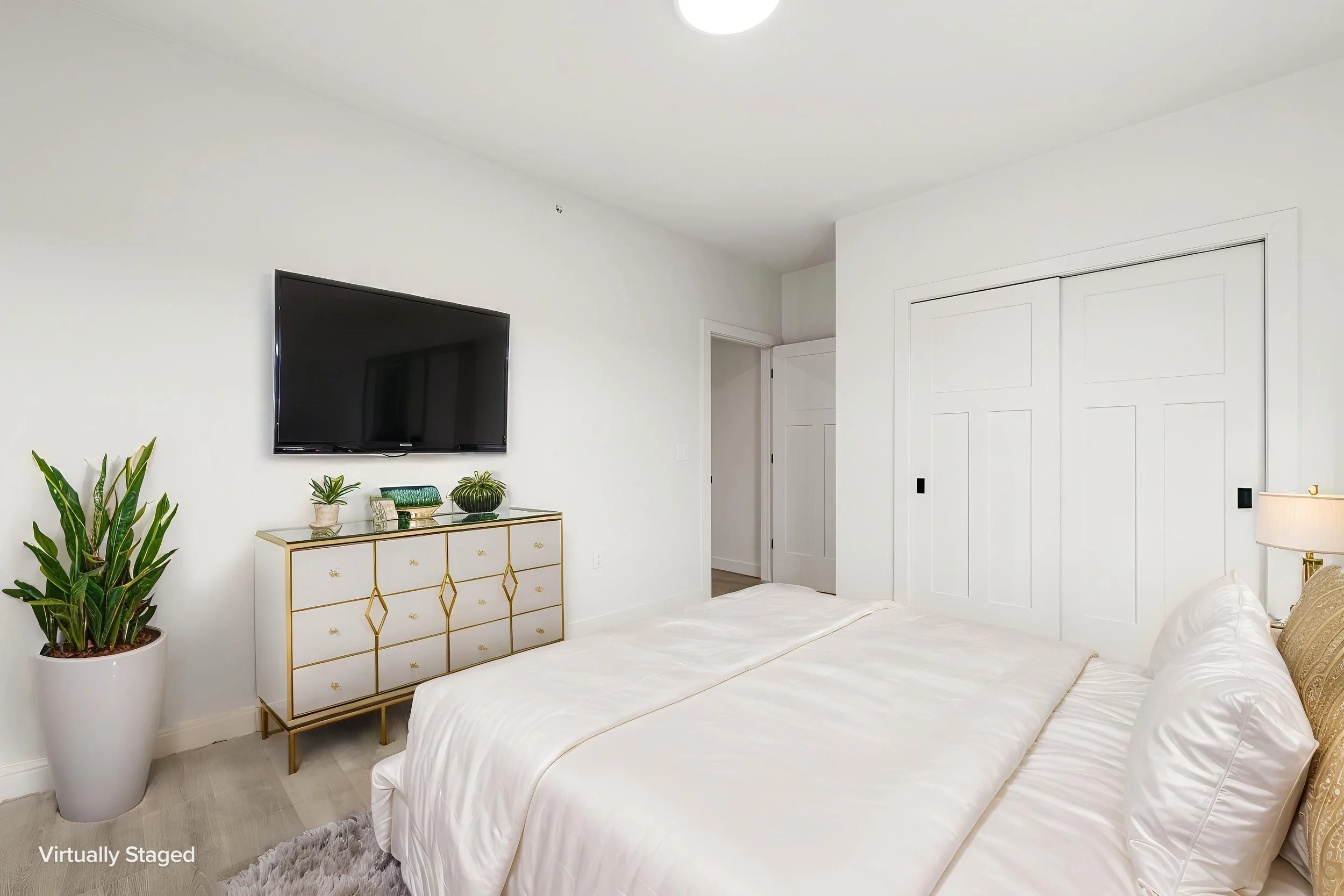 A bedroom with white walls, a large bed with white linens, a gold and white dresser, a potted plant, a wall-mounted TV, and a closet with sliding doors. The Residences at North Harbor on Lake Superior in Marquette Michigan.
