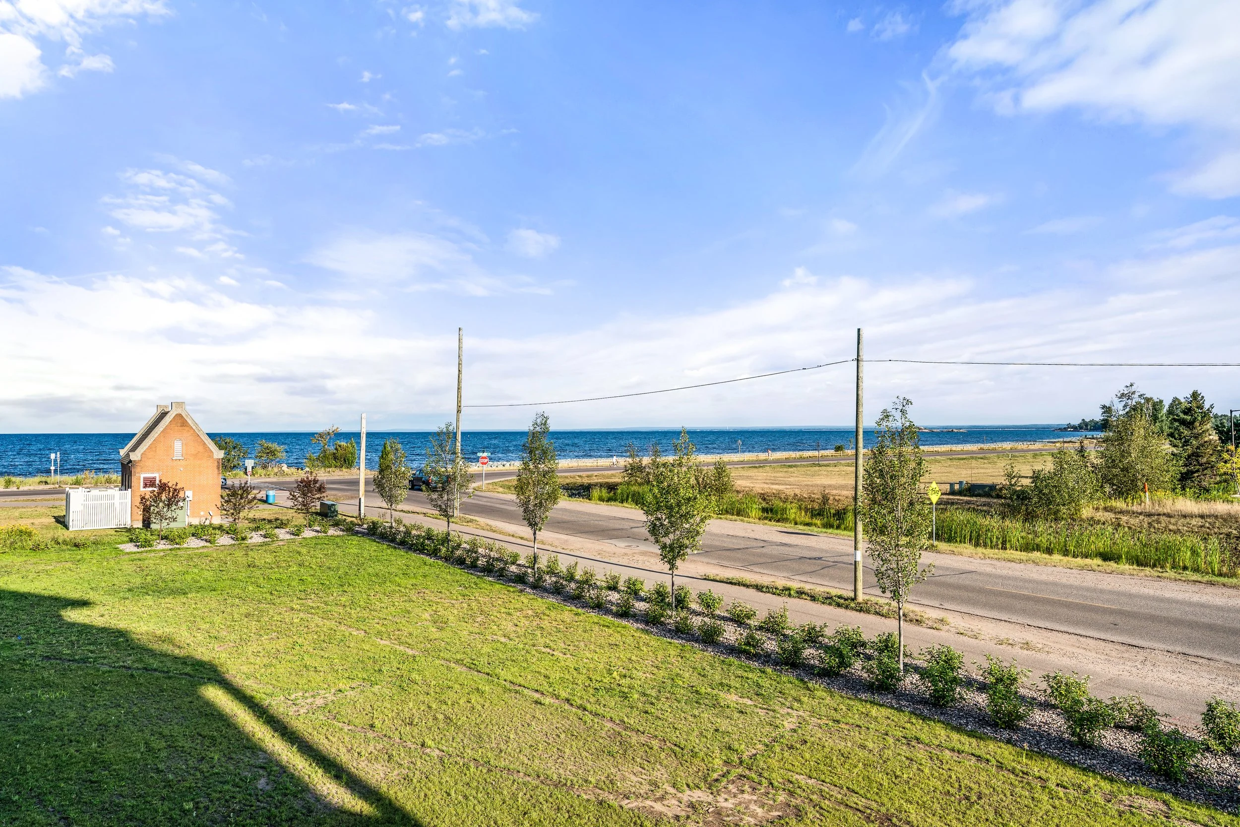 A scenic view of a coastal area with a small brick house, a grassy yard, paved road, trees, utility poles, and the ocean with blue sky and scattered clouds in the background. The Residences at North Harbor on Lake Superior in Marquette Michigan.