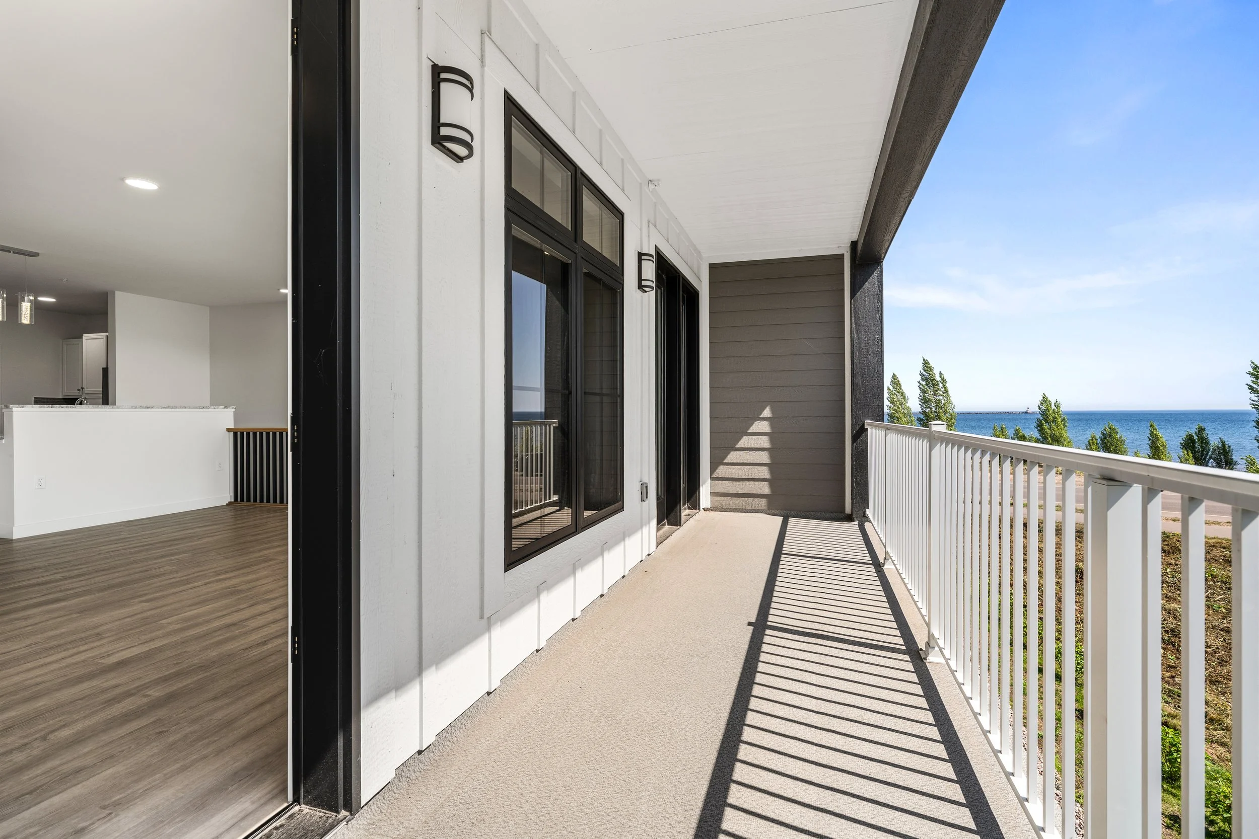 Balcony with white railing, view of trees and ocean in distance, part of modern apartment building with sliding glass door, gray siding, and black accents on the exterior. The Residences at North Harbor on Lake Superior in Marquette Michigan.