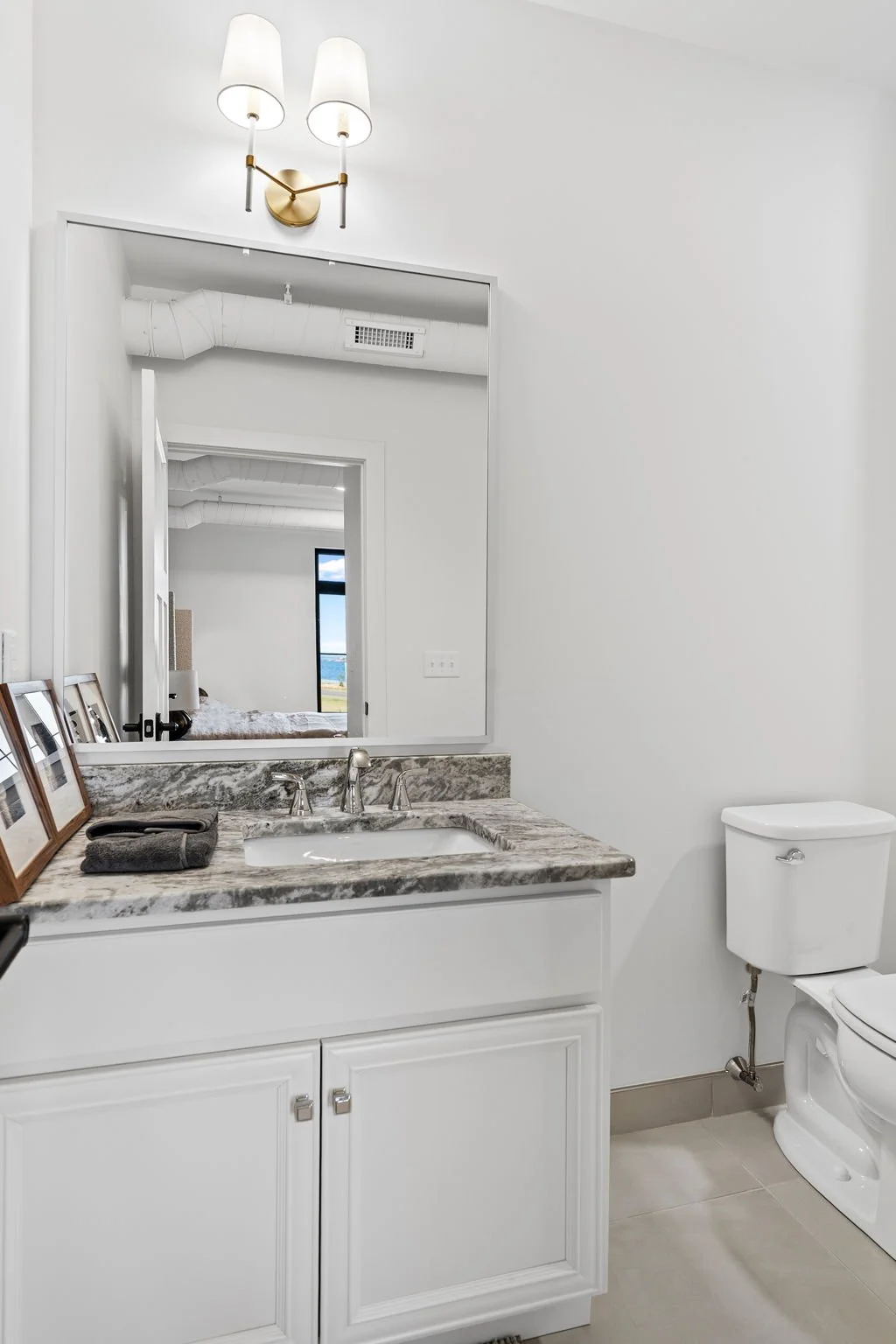 Modern bathroom with white painted walls, a marble countertop vanity, a mirror reflecting a bedroom and window, framed pictures on the counter, a gray towel, and a white toilet. The Residences at North Harbor on Lake Superior in Marquette Michigan.