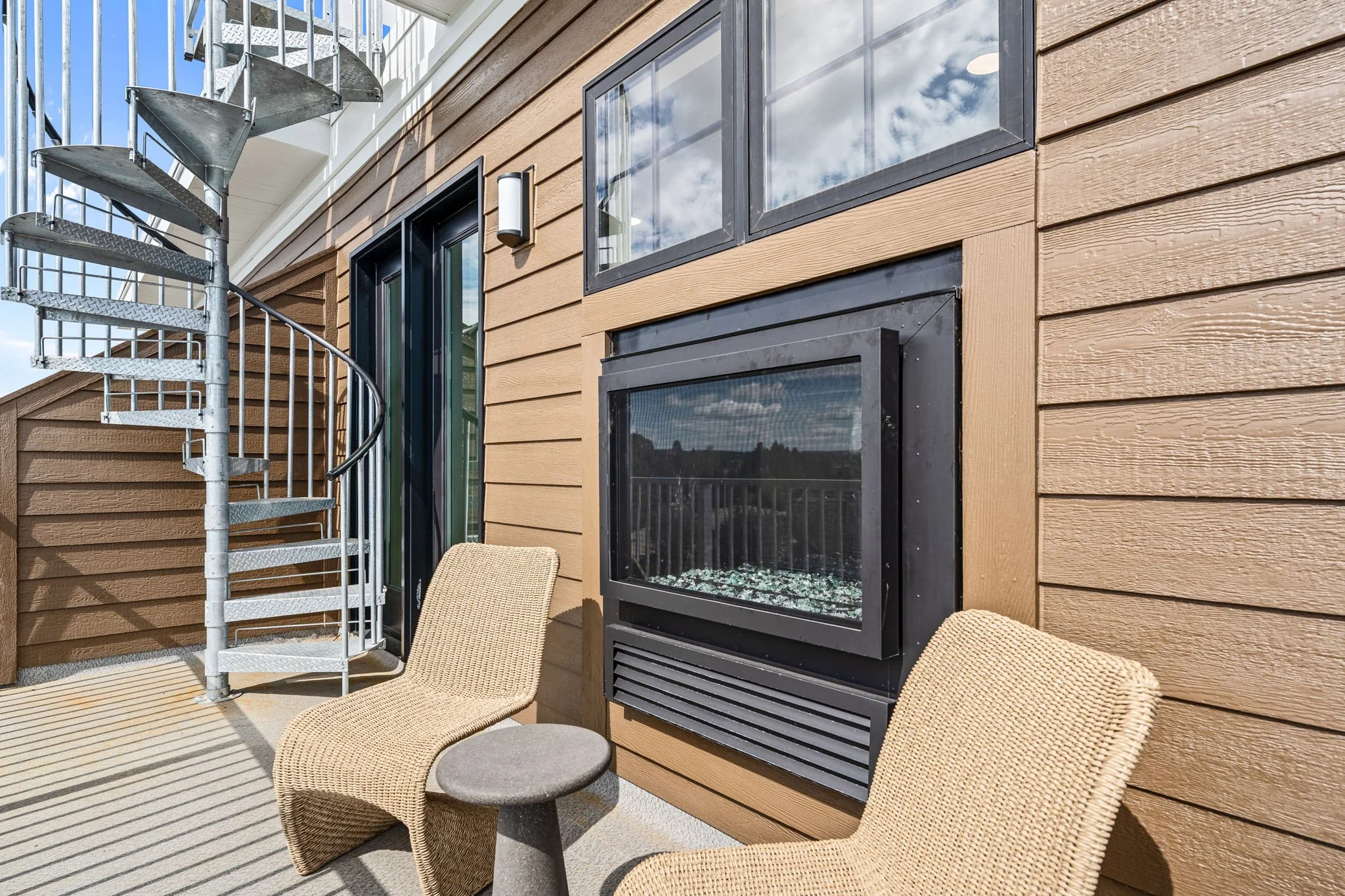Outdoor patio area with beige woven chairs, a small dark table, a decorative fire feature, a metal spiral staircase large windows. The Residences at North Harbor on Lake Superior in Marquette Michigan.