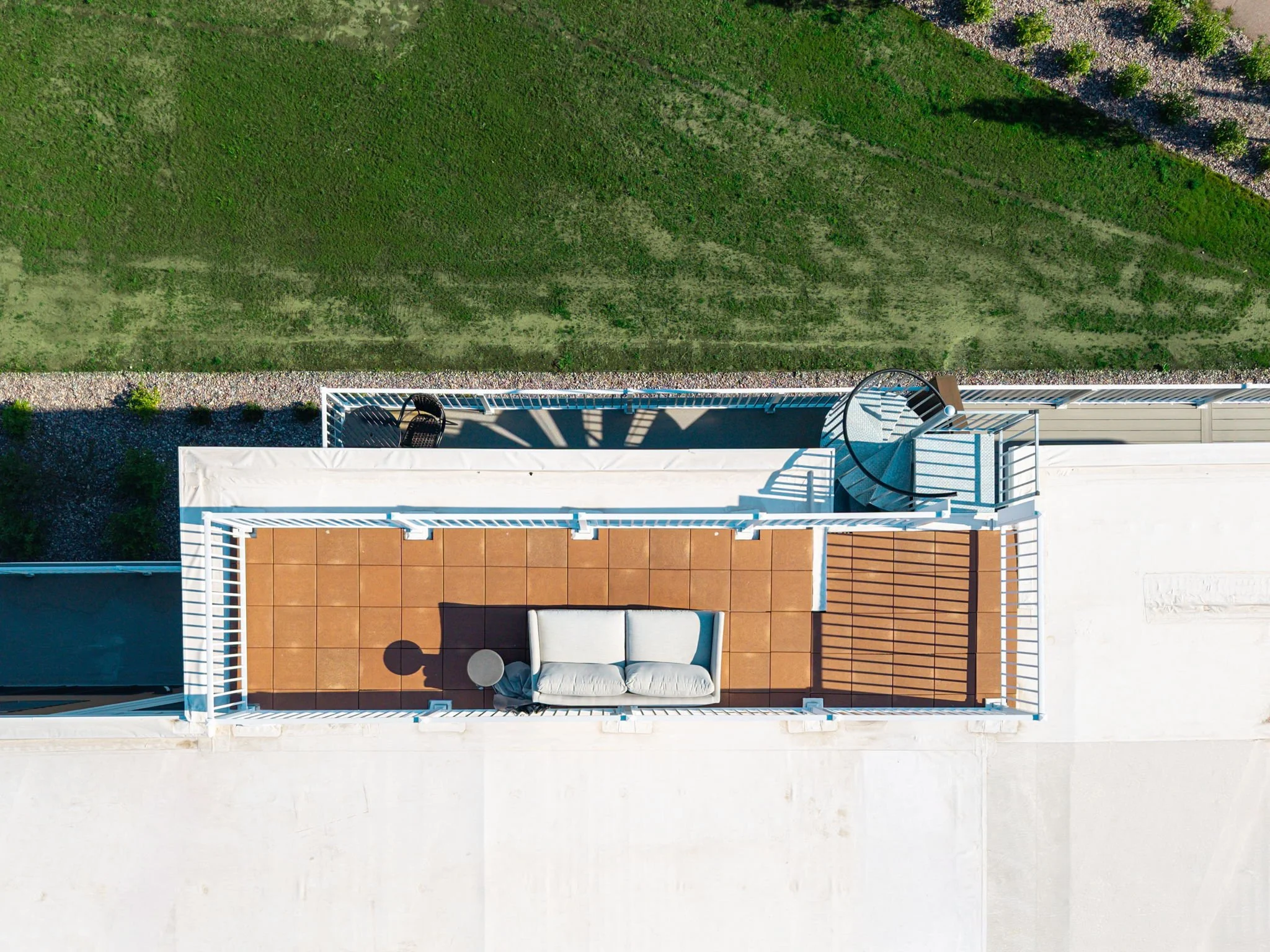 Aerial view of a balcony with a white railing, outdoor furniture including a curved sofa with white cushions, small side tables, and a terracotta-tiled floor. The Residences at North Harbor on Lake Superior in Marquette Michigan. 