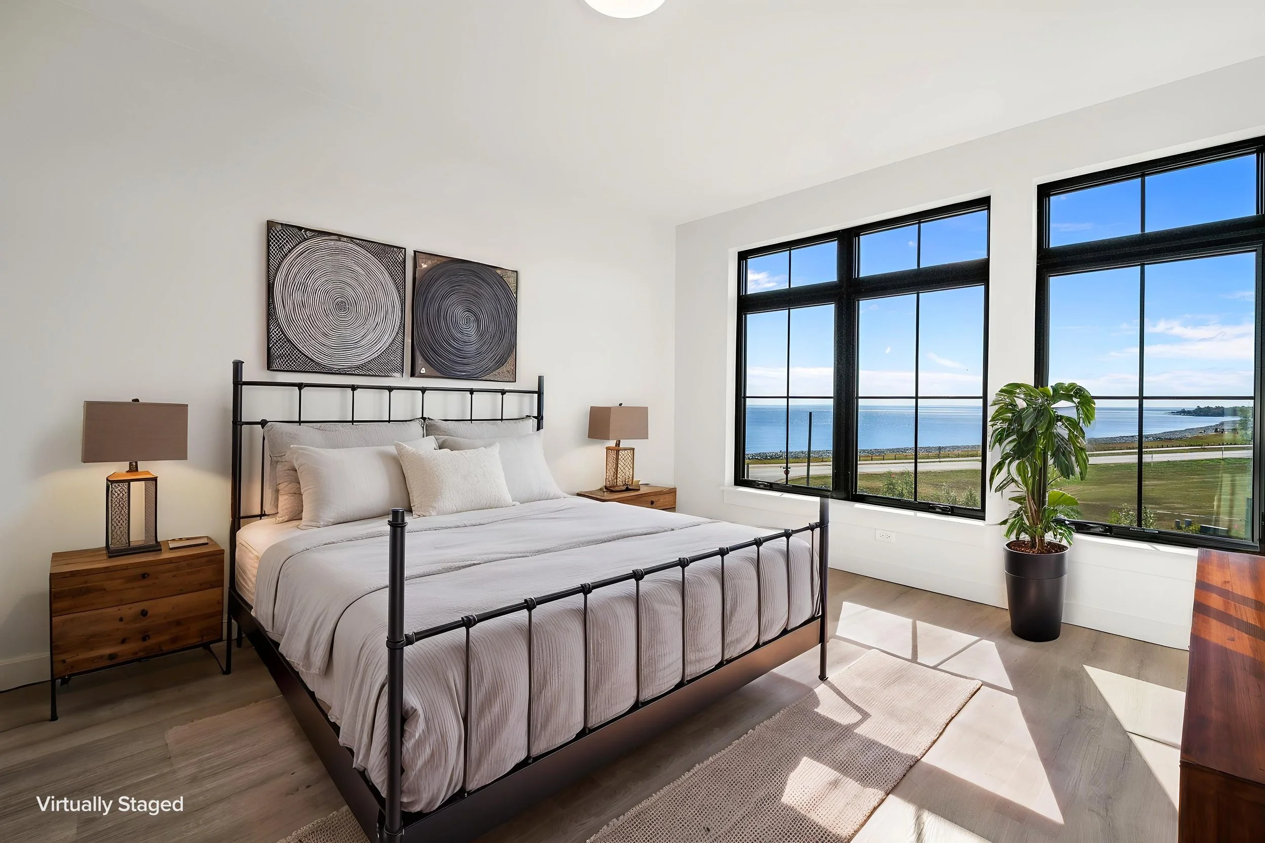 A bedroom with a metal bed frame, two wooden nightstands with lamps, artwork above the bed, large windows with a view of water and sky, and a potted plant. The Residences at North Harbor on Lake Superior in Marquette Michigan.