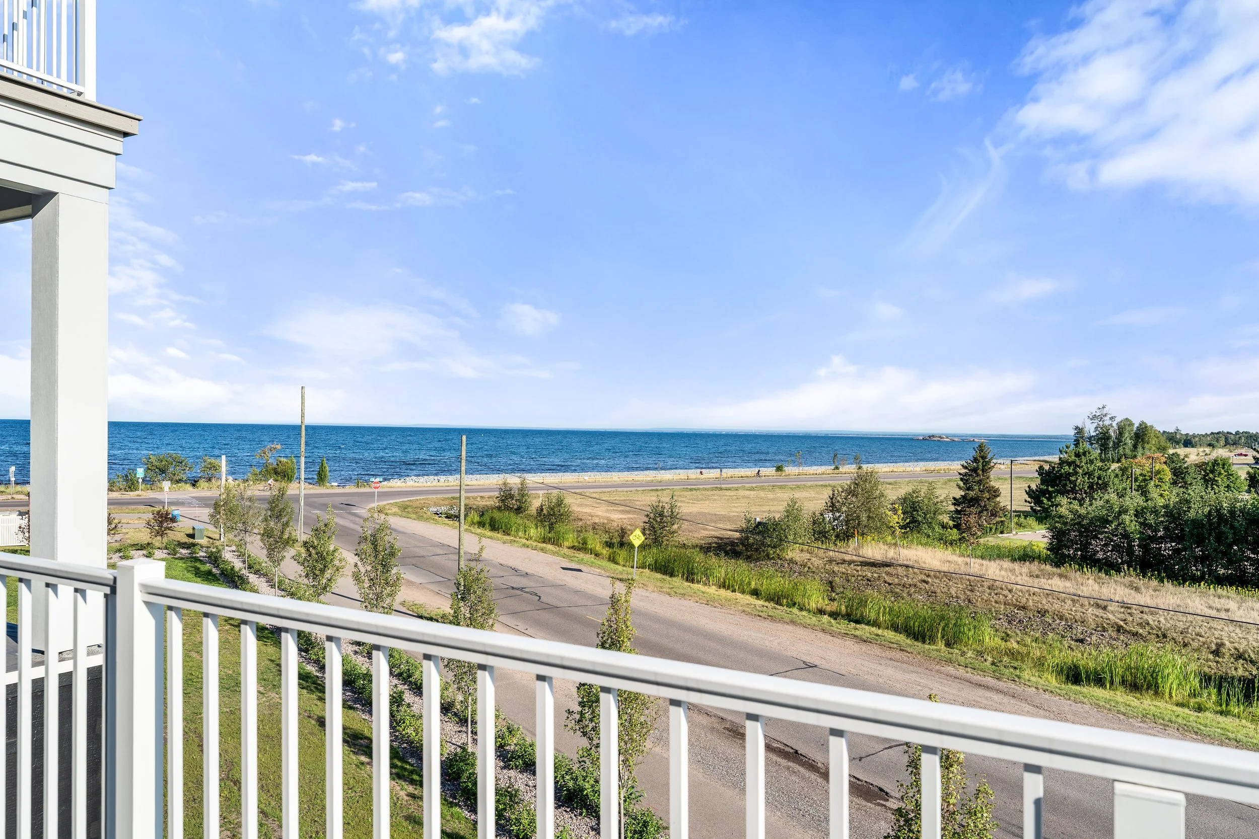View of a coastline with a blue ocean, a partly cloudy sky, a road, and green grass and trees from a balcony with a white railing. The Residences at North Harbor on Lake Superior in Marquette Michigan.