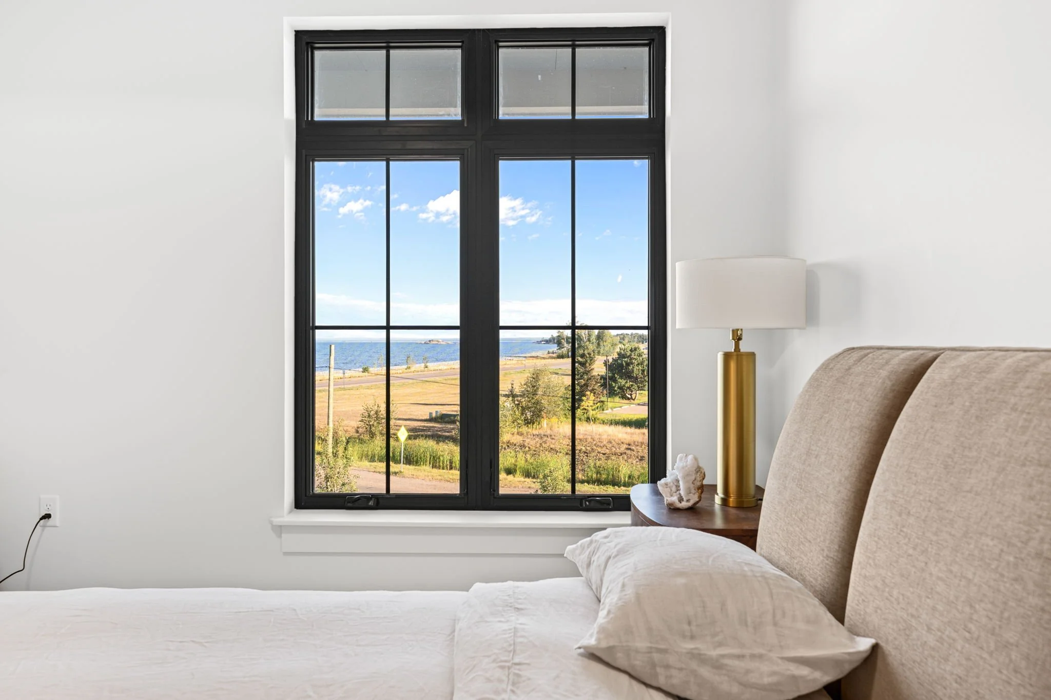 A bedroom with a large black-framed window revealing a view of a coastal landscape with blue sky, clouds, water, and greenery. The Residences at North Harbor on Lake Superior in Marquette Michigan.