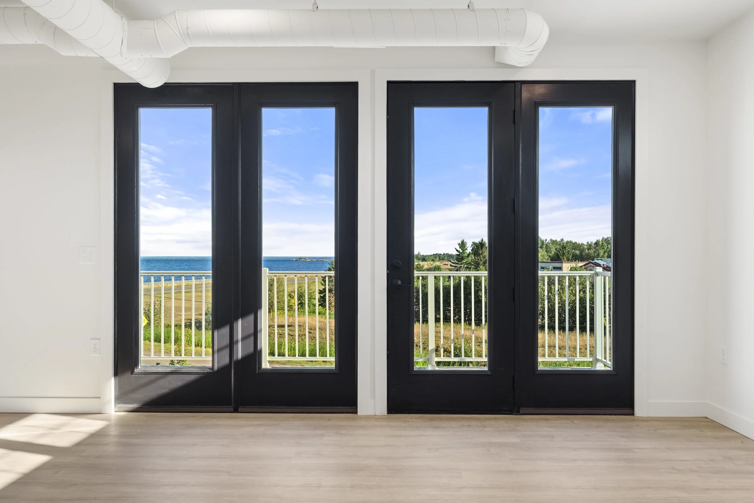 Interior view of a room with double black-framed glass doors leading to a balcony, showing an ocean view with a blue sky and greenery beyond. The Residences at North Harbor on Lake Superior in Marquette Michigan.