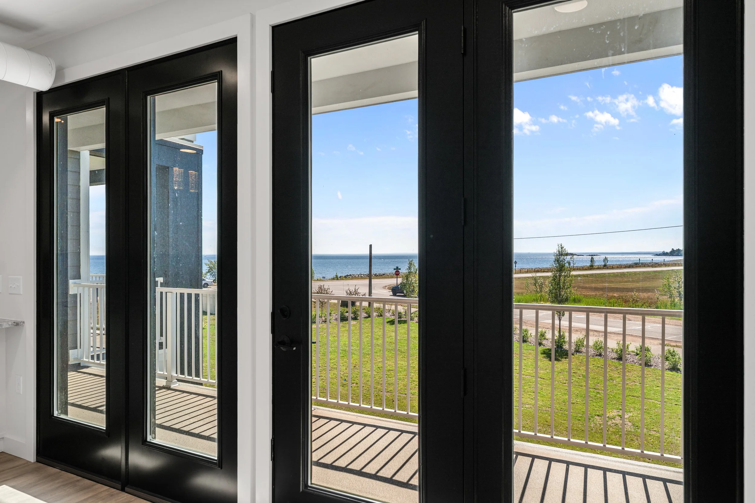 Interior view of black-framed glass sliding doors opening to a balcony with a view of the ocean, blue sky with clouds, and a grassy area with a sidewalk outside. The Residences at North Harbor on Lake Superior in Marquette Michigan.
