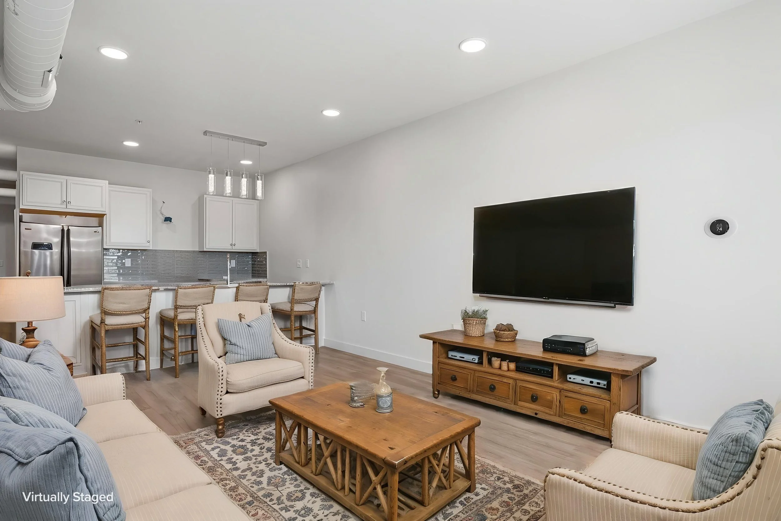 Living room with beige sofas and chairs, wooden coffee table, TV mounted on white wall, kitchen area in background, hardwood floors, decorative pillows and plants. The Residences at North Harbor on Lake Superior in Marquette Michigan.