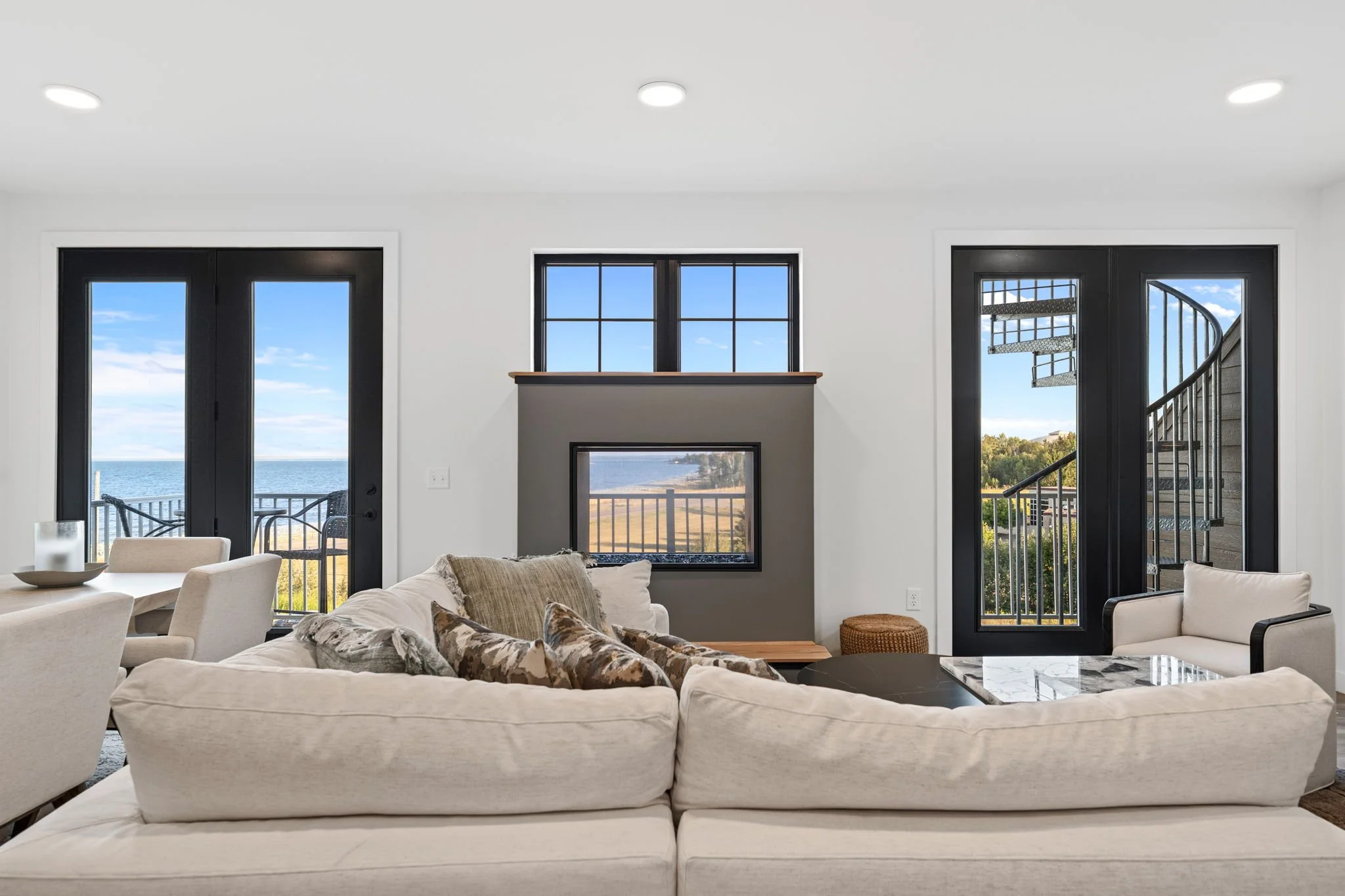 Living room with large windows and a view of the coastline, featuring a white sectional sofa, a beige armchair, and sliding glass doors leading to an outdoor balcony. The Residences at North Harbor on Lake Superior in Marquette Michigan.