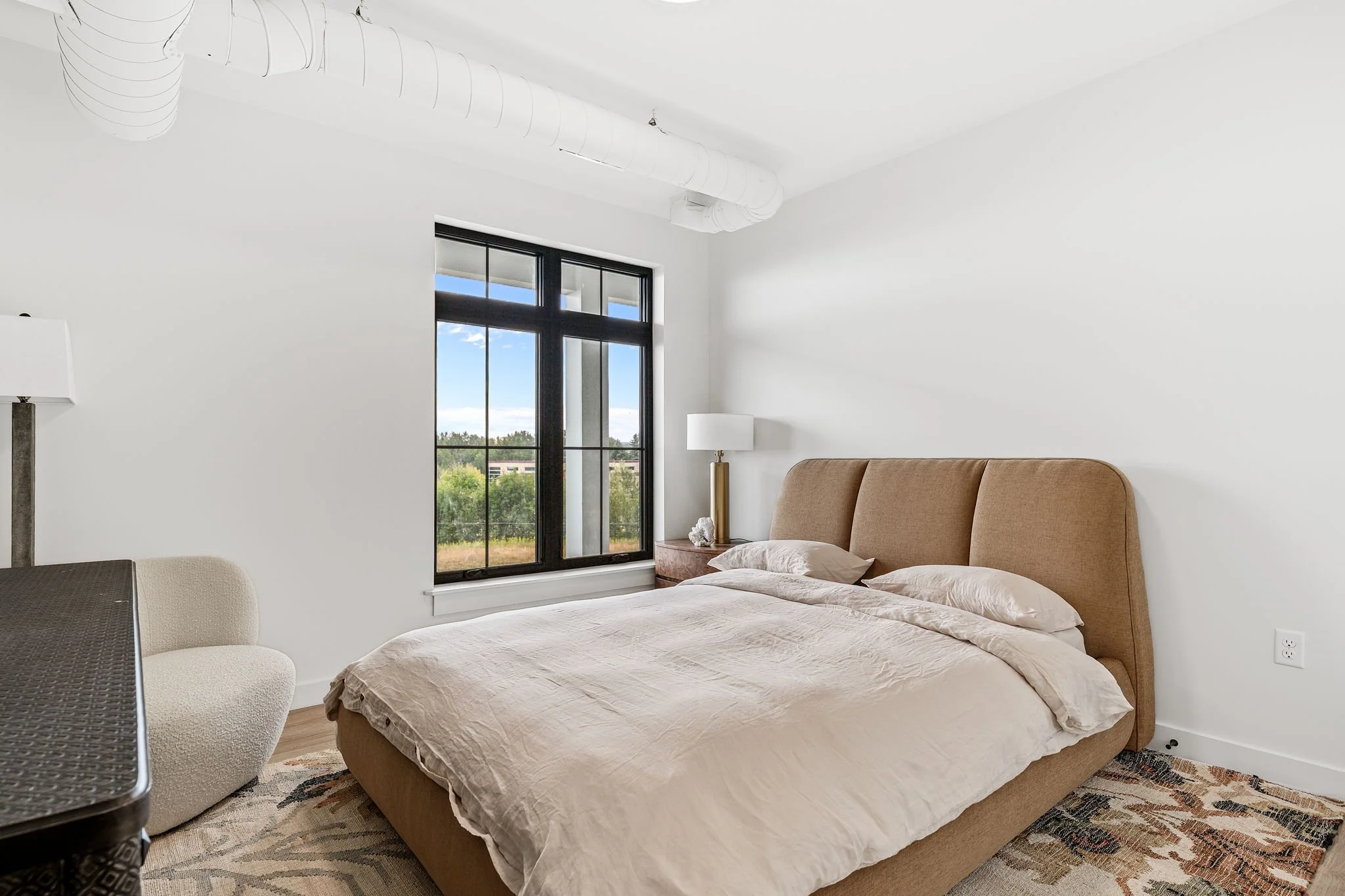 Bedroom with beige upholstered bed, white bedsheets, and pillows. Natural light coming through a large window with black frames, showing greenery outside. The Residences at North Harbor on Lake Superior in Marquette Michigan.