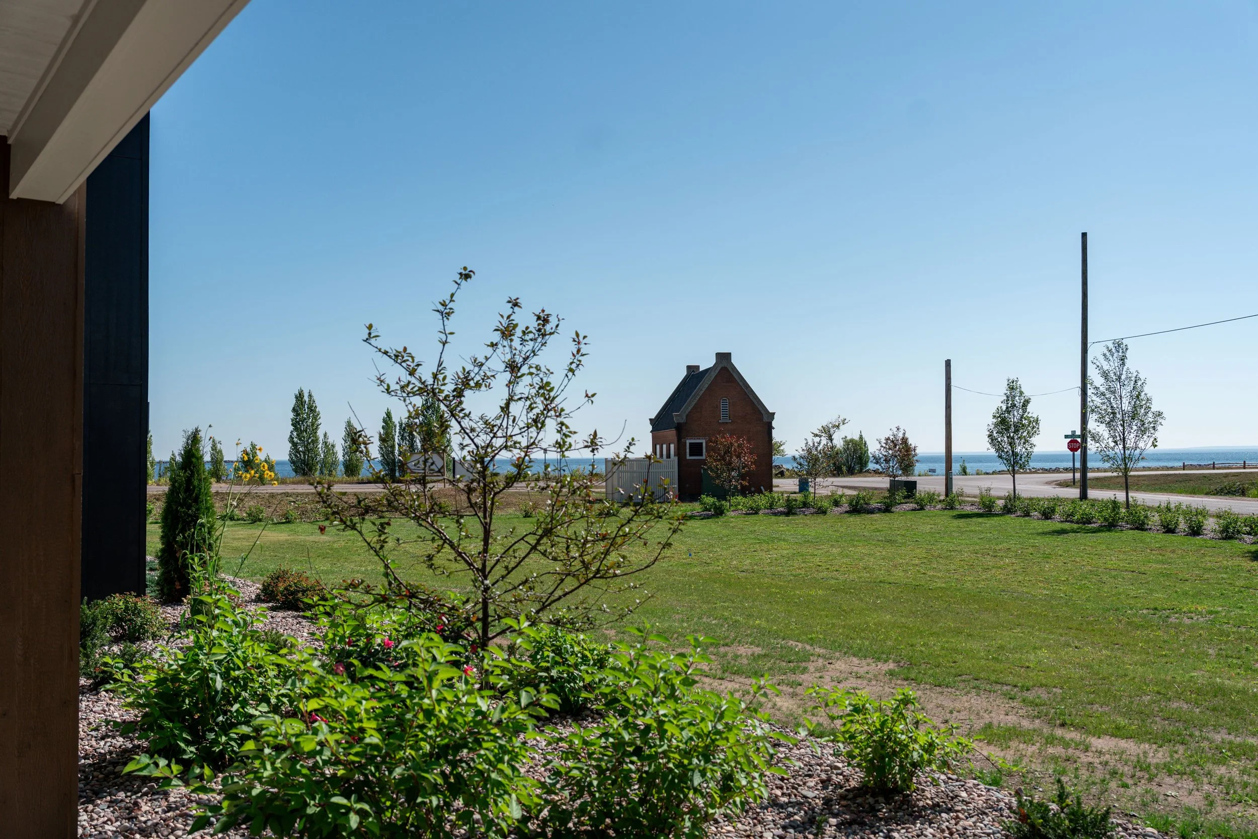 View of a backyard garden with green grass, flowering plants, and trees, overlooking a street with utility poles and a small brick building near the water in the distance. The Residences at North Harbor on Lake Superior in Marquette Michigan.